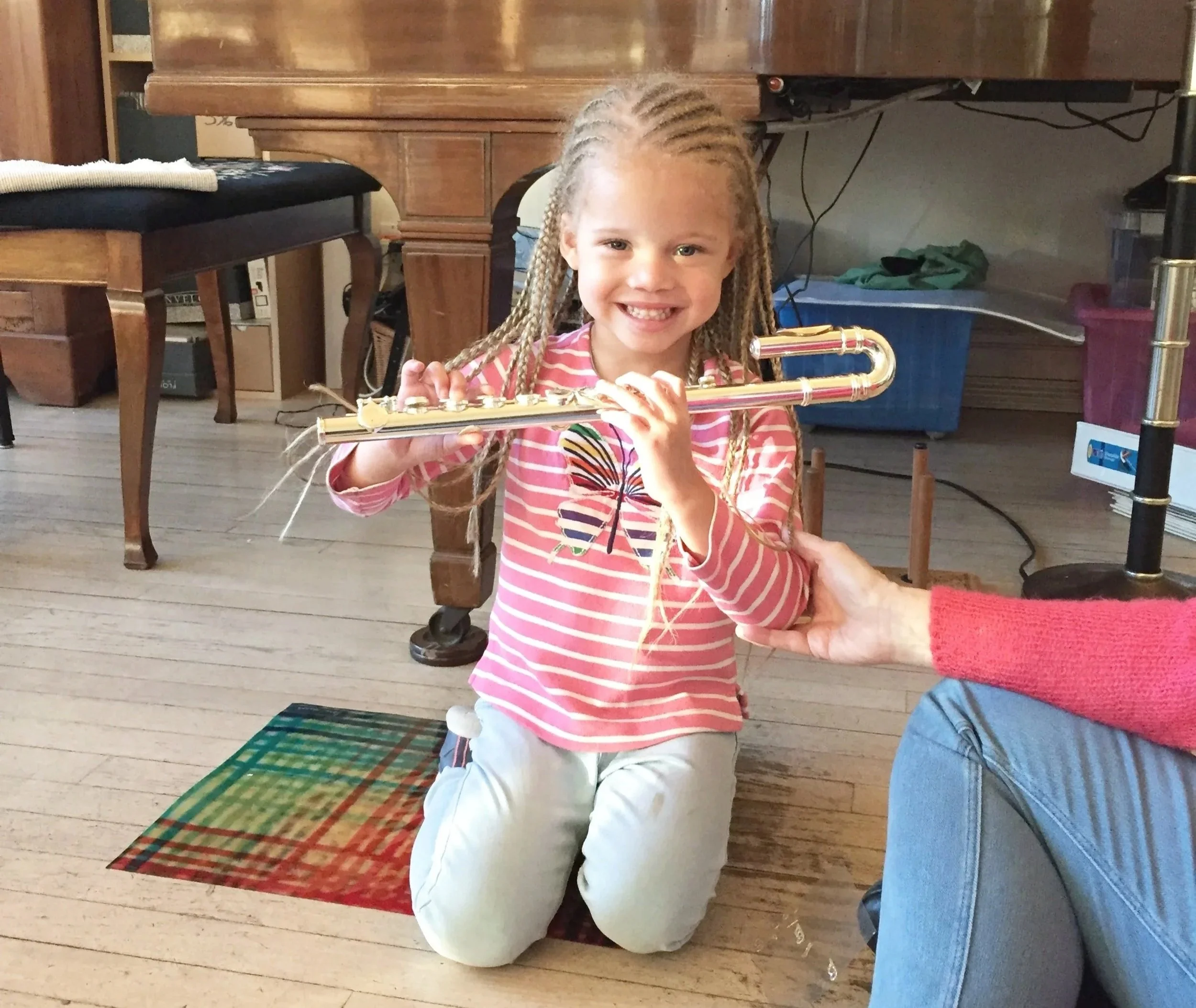 One of Wendy's Suzuki flute students, kneeling on the floor and holding a curved headjoint flute and smiling. Caregivers's hand is supporting her elbow