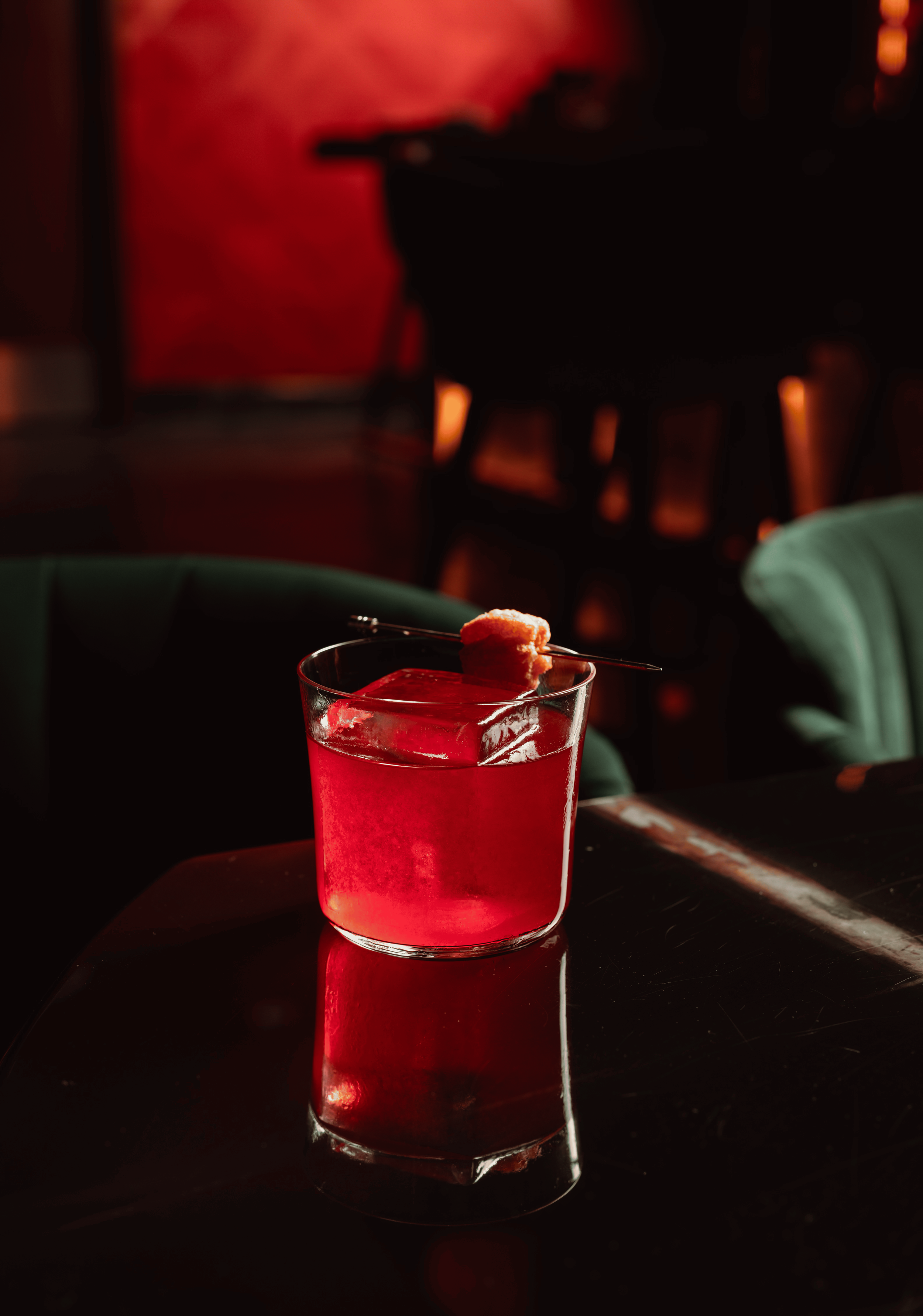 A vibrant red craft cocktail served in a rocks glass with a single large, clear ice cube and an orange peel garnish on a dark, reflective table.