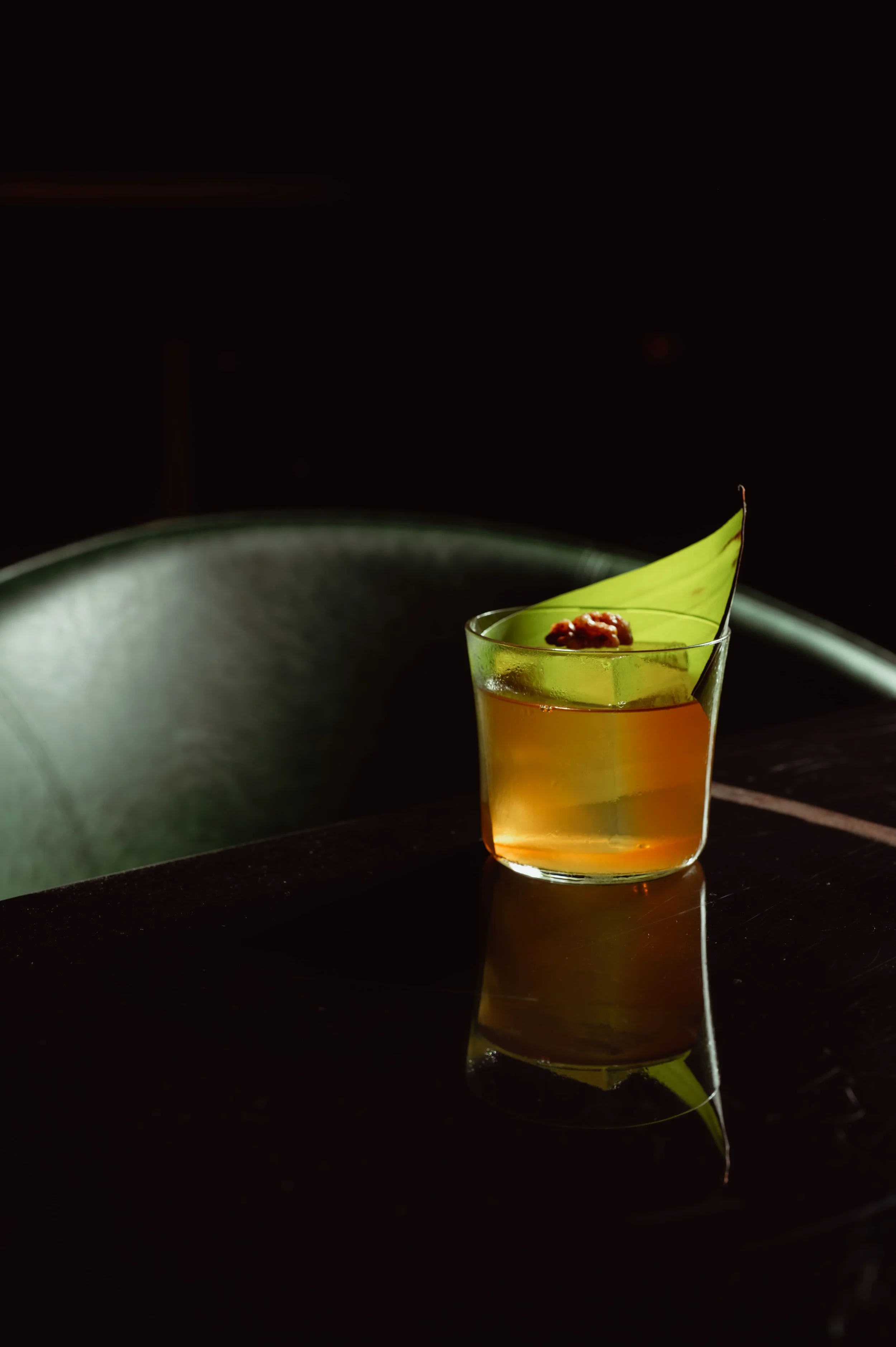 An amber-colored cocktail on a dark table, garnished with a large green leaf and a candied nut, set against a moody, dark green interior.