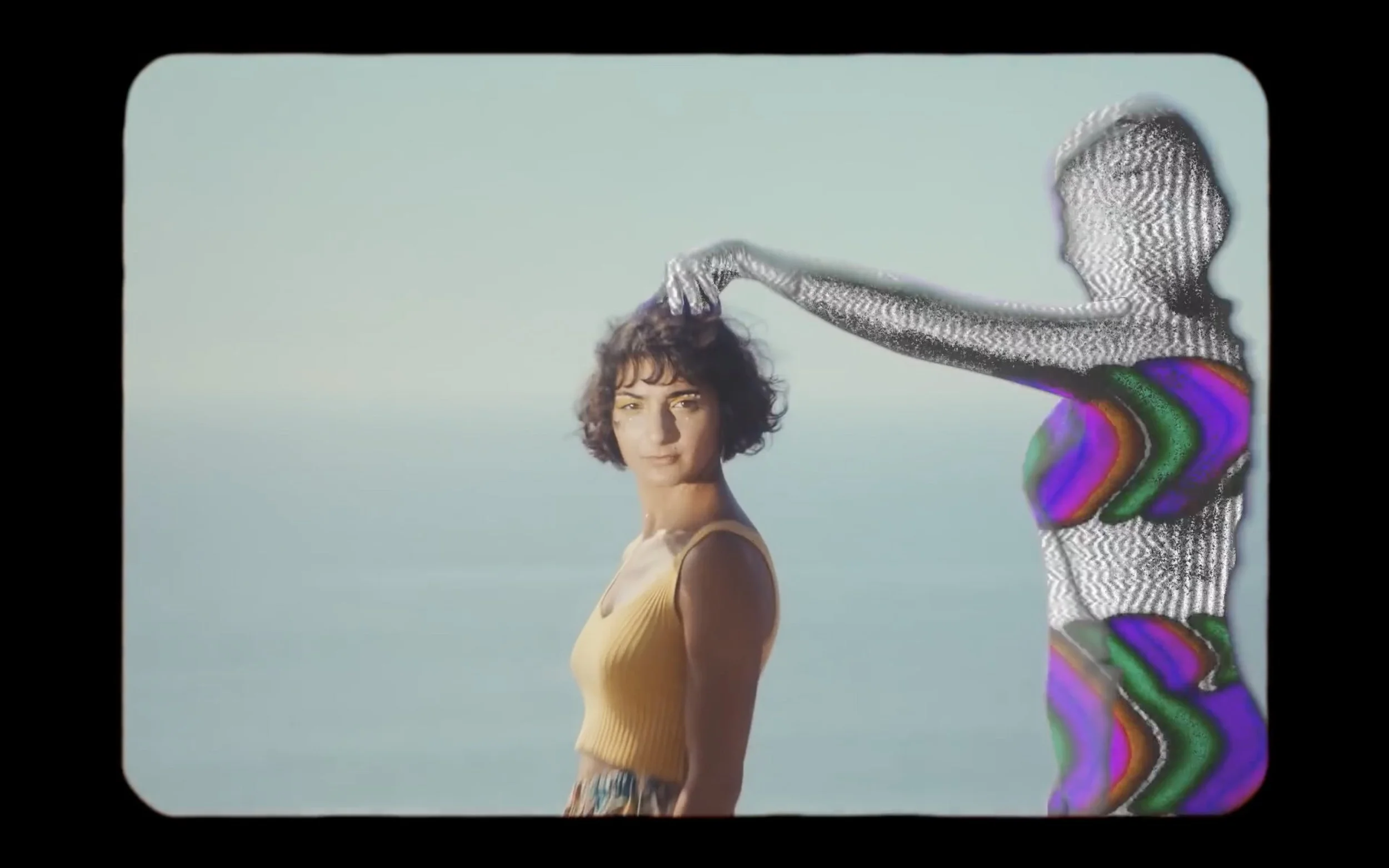 A woman in a yellow tank top posing outdoors with a neutral expression, while a digital figure in black and white with vibrant, colorful accents on the chest and waist interacts with her by touching her hair.