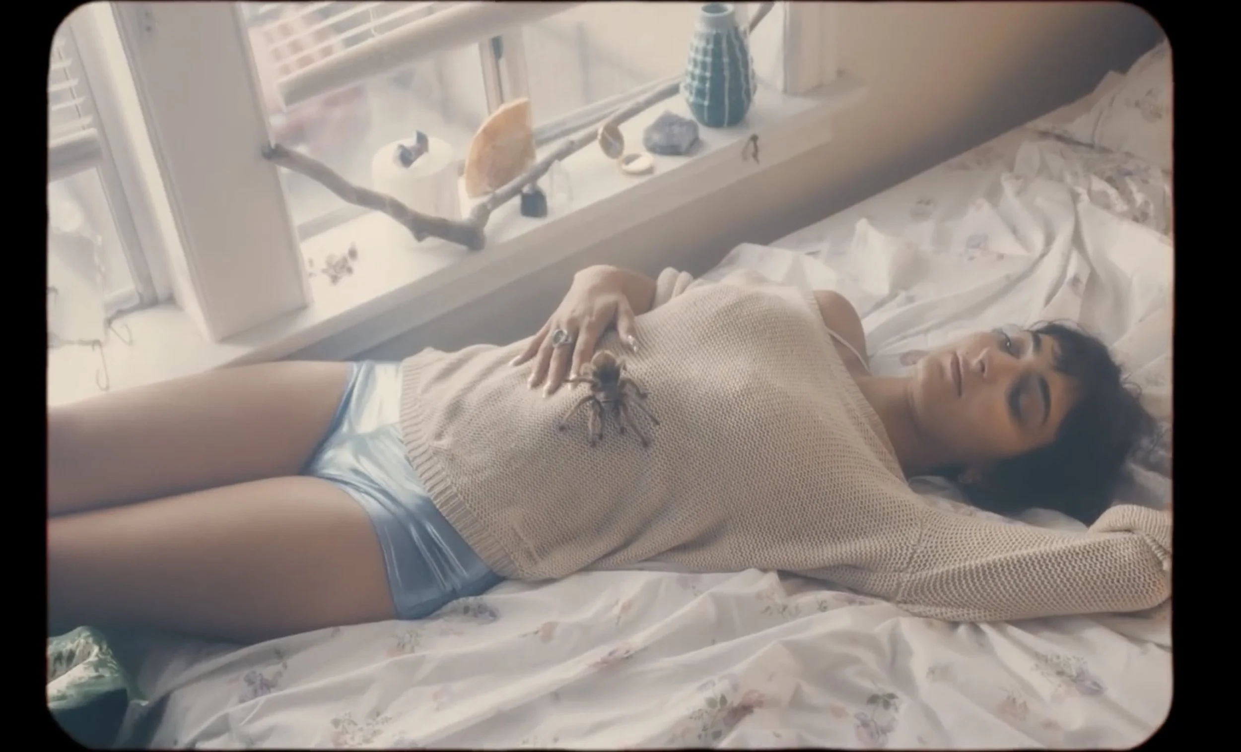 A woman lying on a bed with a tarantula on her chest, with a window and decorative objects on the windowsill in the background.