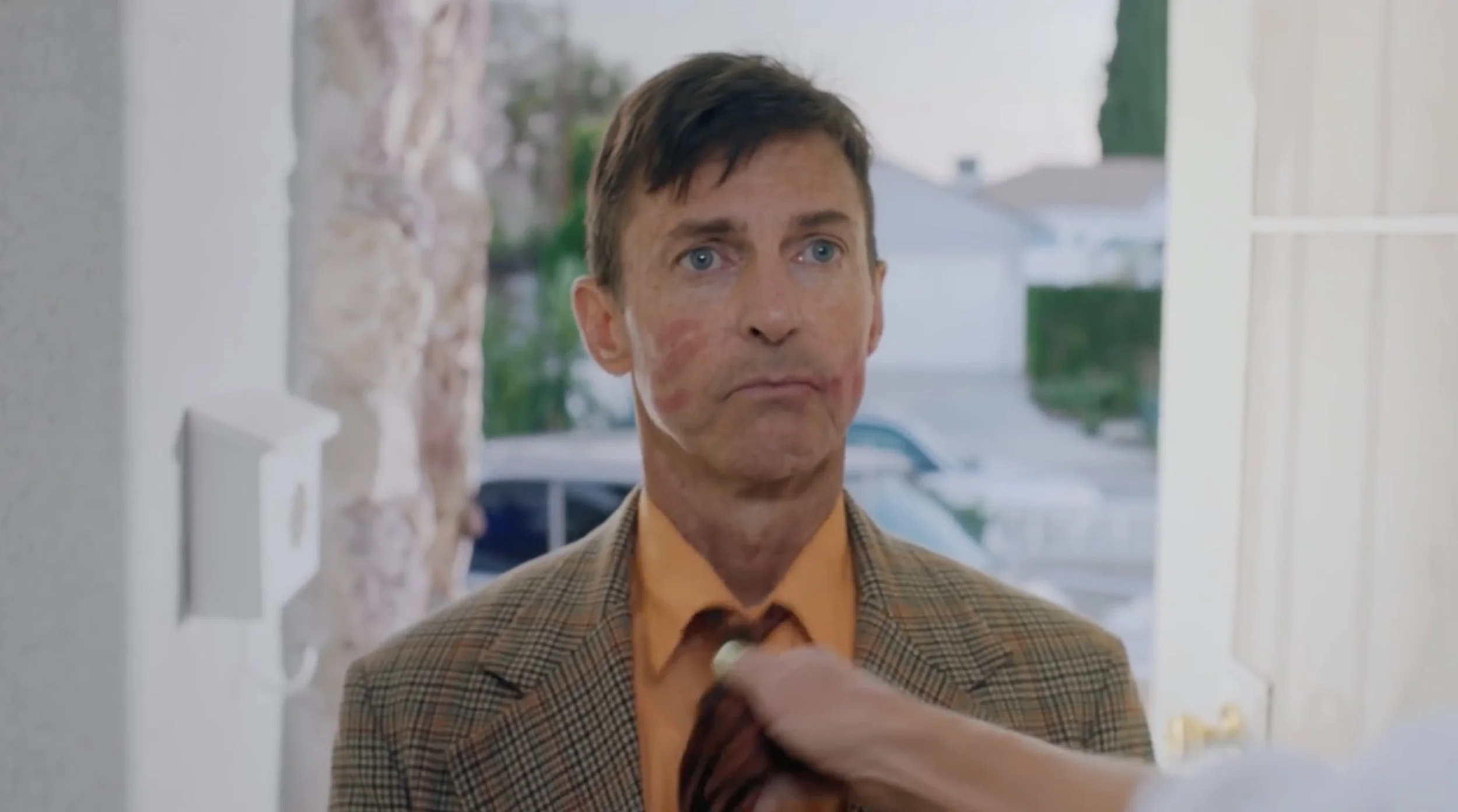 A man with a blank expression standing at a doorway, with a hand wiping or smearing his face, which has red marks or makeup.