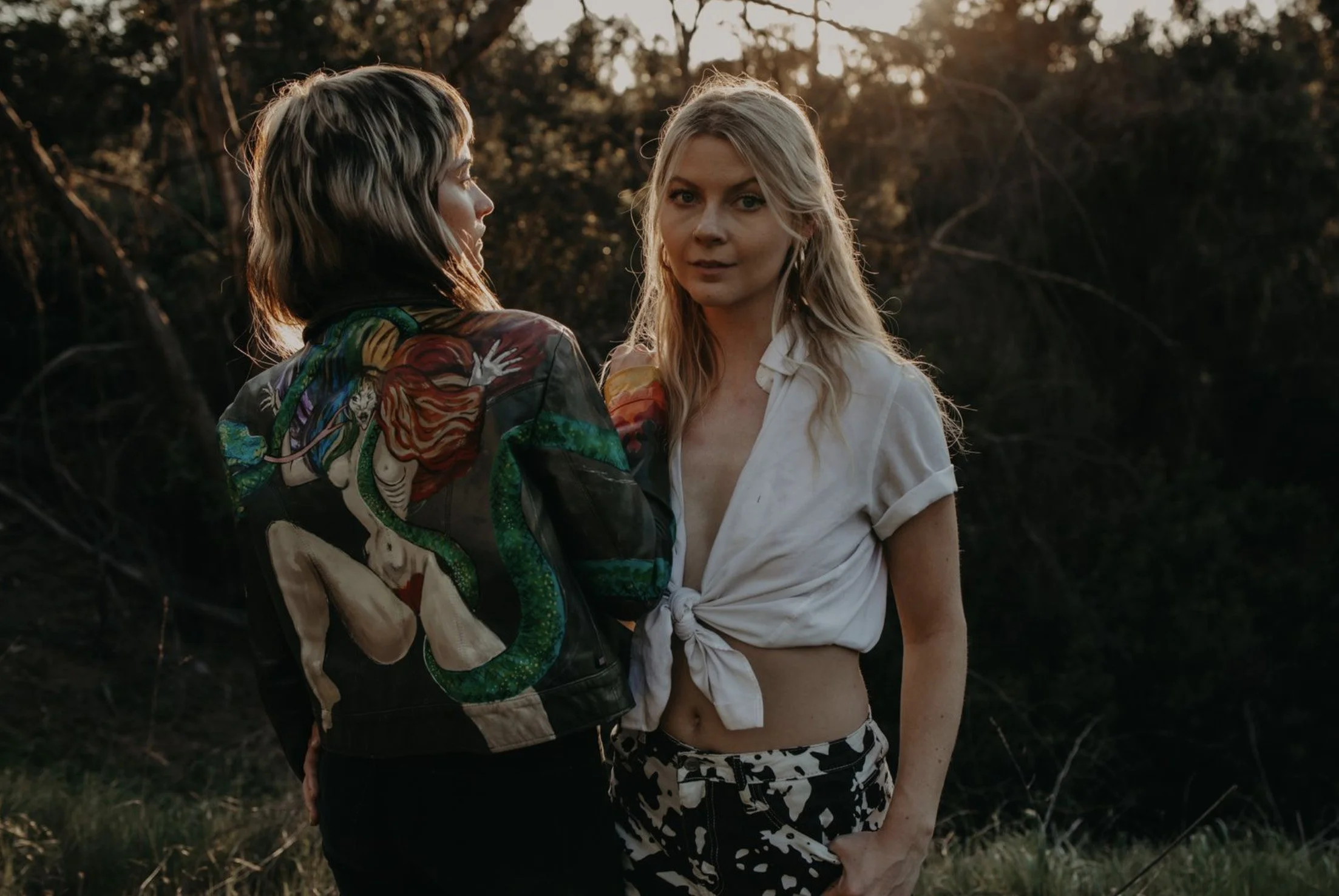Two women standing outdoors during sunset, one facing sideways and the other facing forward. The woman on the left has short, dark hair and is wearing a colorful jacket with artistic designs. The woman on the right has long, blonde hair and is wearin