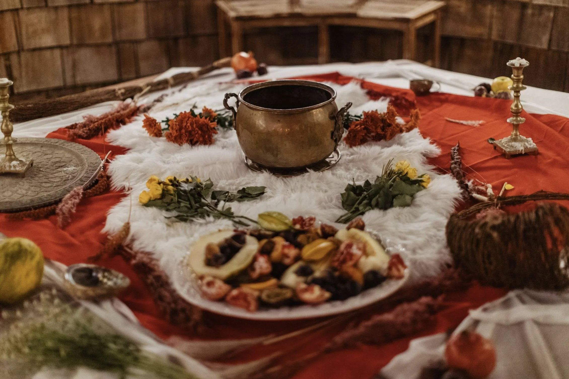 A rustic table set with a brass pot, flowers, candles, and a variety of food, including a platter of assorted foods, against a wooden wall background.
