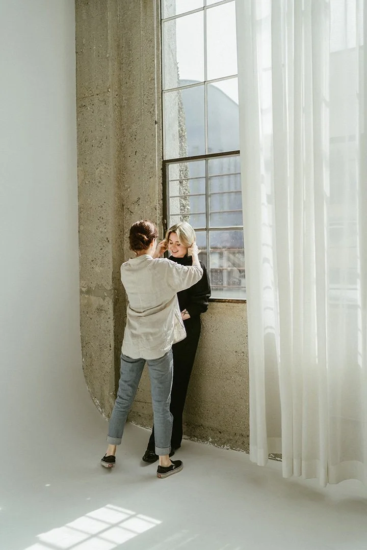 Two women in a brightly lit room near a large industrial-style window with sheer white curtains. One woman is touching the other's face, and they are smiling at each other.
