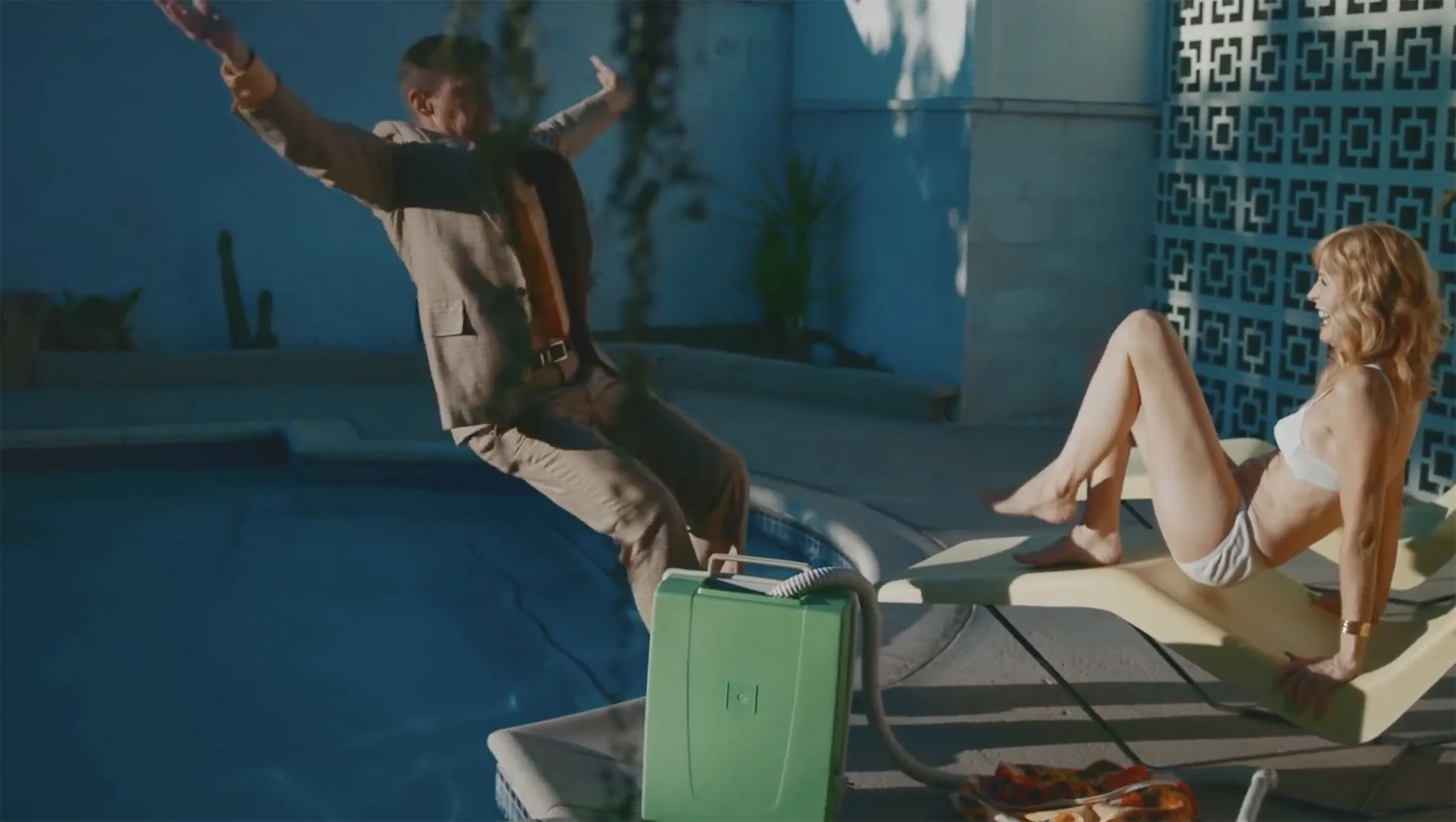 A man and woman near a swimming pool at night, with the man in mid-fall from the pool edge and the woman lounging on a poolside chair, smiling.