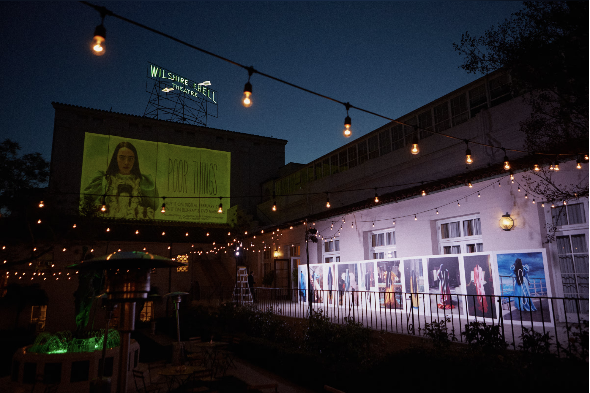 Nighttime view of an outdoor art exhibit with string lights, a pink building with large windows, and a large screen showing a woman and the words 'POOR THINGS.' There are framed artworks displayed along the building's wall.