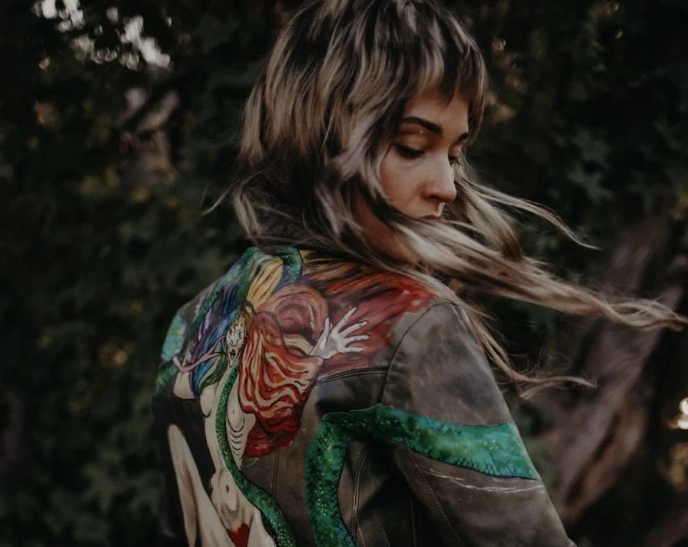 A woman with long hair looking downward, wearing a jacket with a colorful, artistic dragon design on the back, outdoors with dark, blurry foliage in the background.