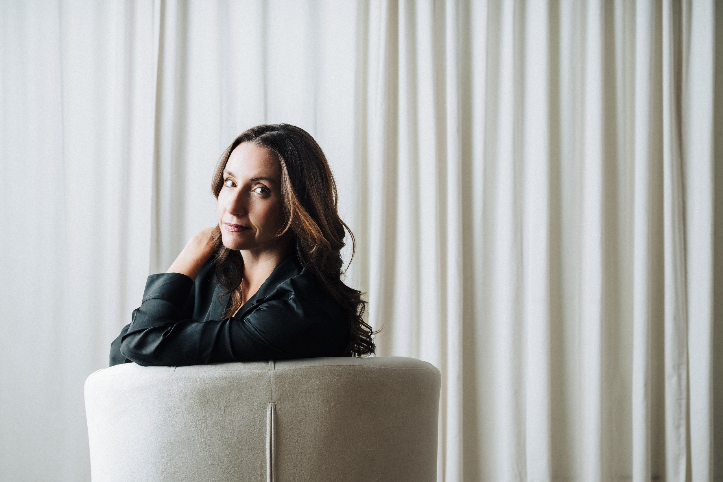 A woman with long brown hair sitting on a white chair in front of a cream-colored curtain, wearing a black leather jacket and gazing at the camera.