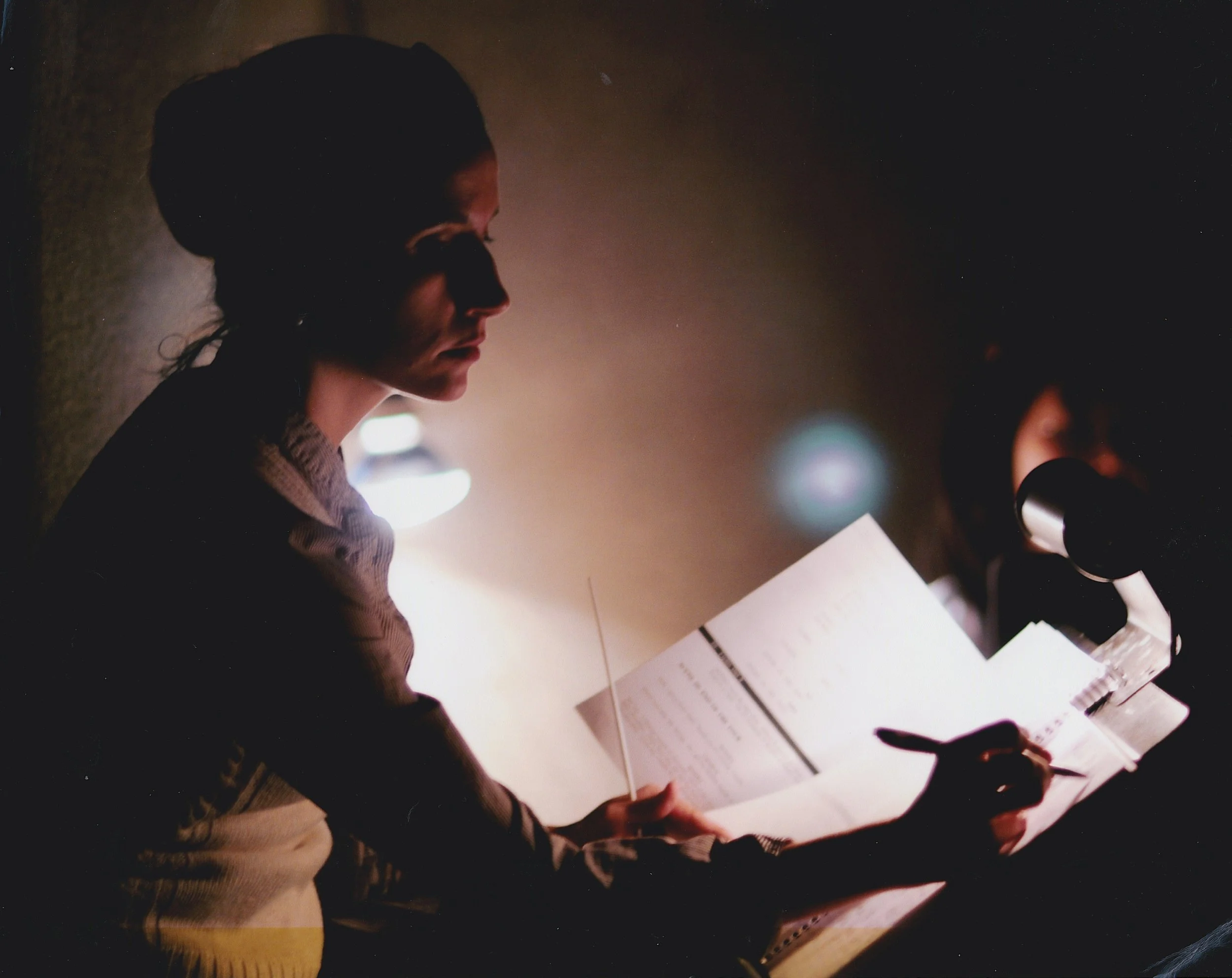 A woman with dark hair tied back, reading a paper and speaking into a microphone in a dimly lit room.