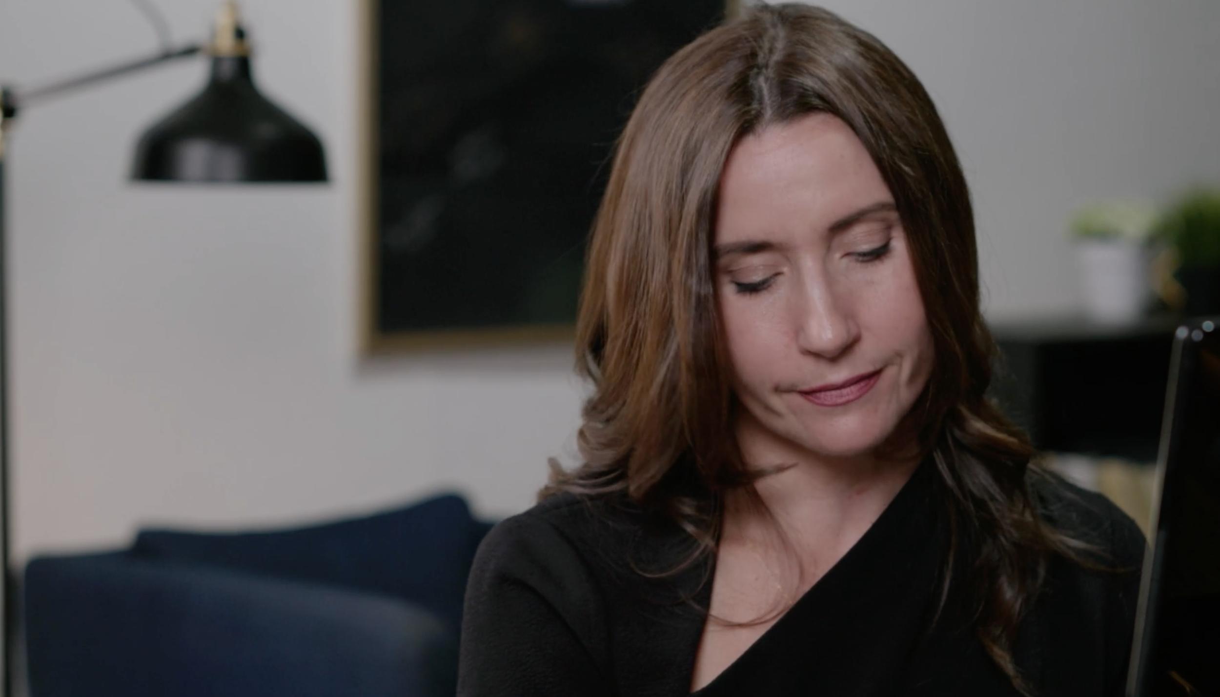 A woman with shoulder-length brown hair looking down with a slight smile, sitting on a dark blue sofa in a modern living room with a black lamp and a piece of wall art in the background.