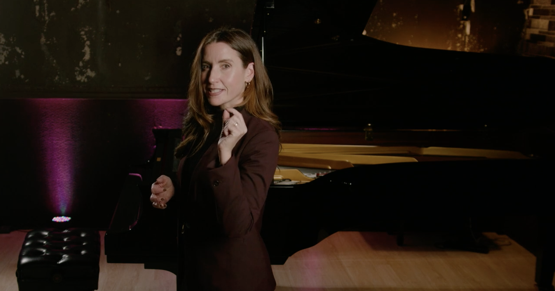 A woman with shoulder-length brown hair stands next to a black grand piano in a dimly lit room with pink and gold lighting accents.