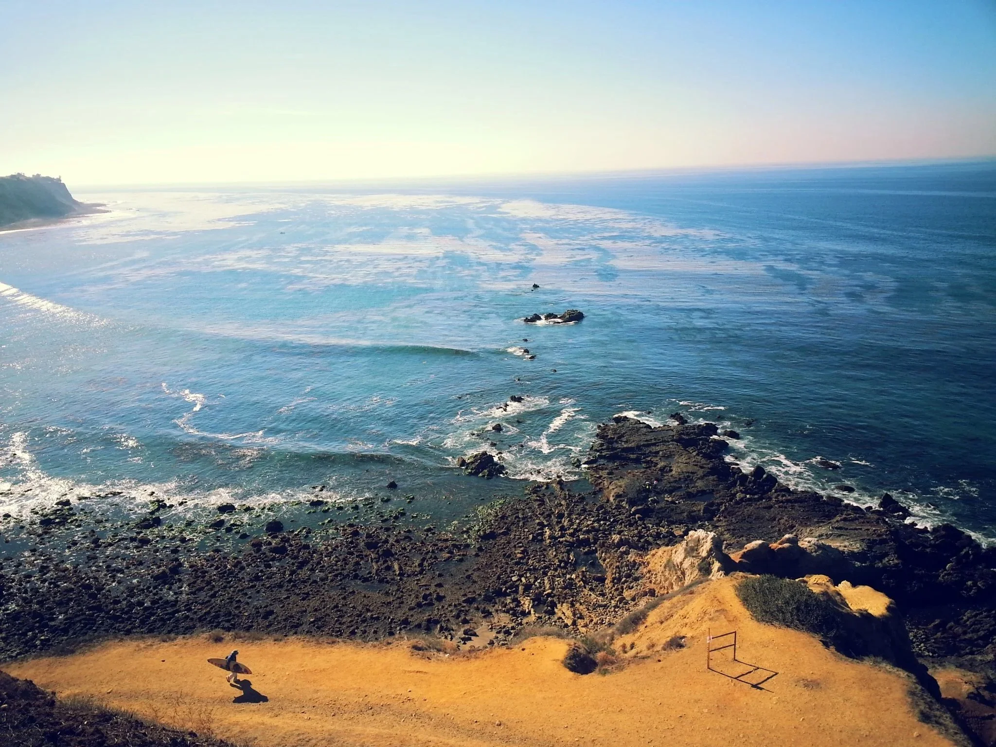 A coastline with rocks and cliffs, ocean waves, and a person carrying a surfboard walking on a sandy path.