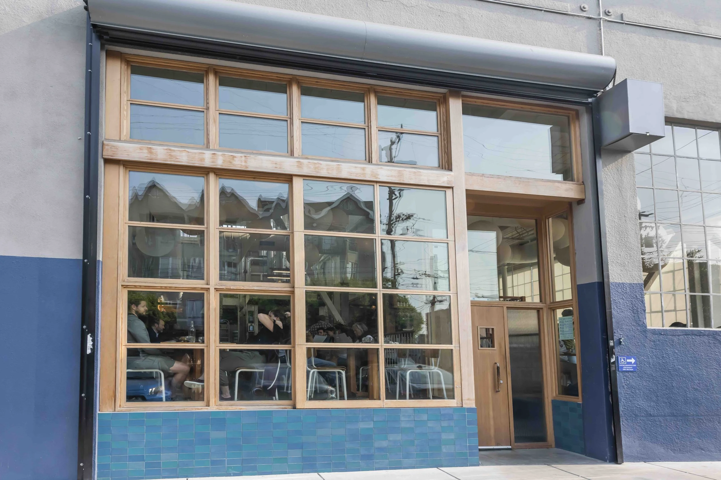 Front of a modern restaurant with large glass windows, wood framing, and blue tiled base, showing people dining inside.