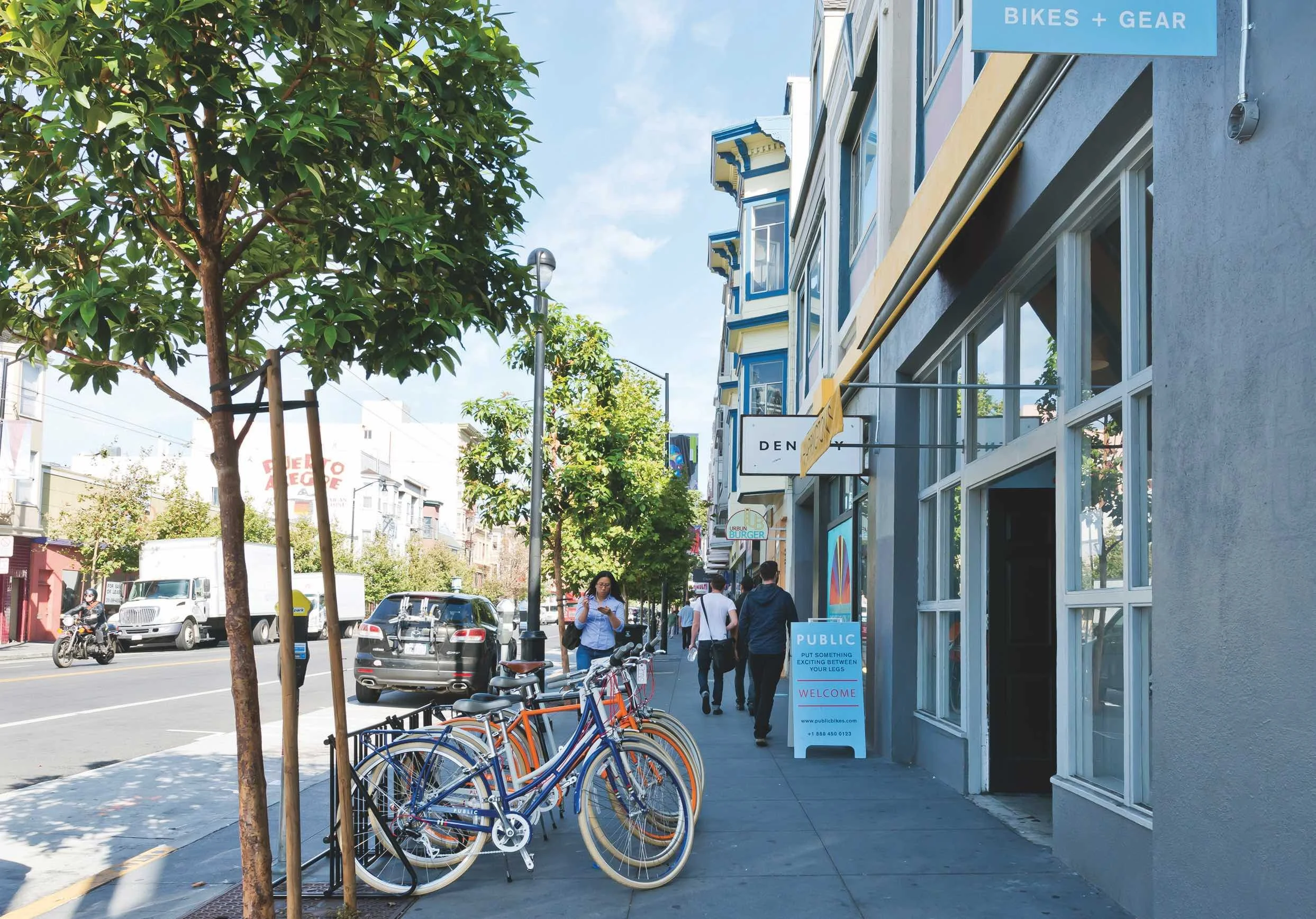 City sidewalk with parked bikes, pedestrians, storefronts, and trees on a sunny day.