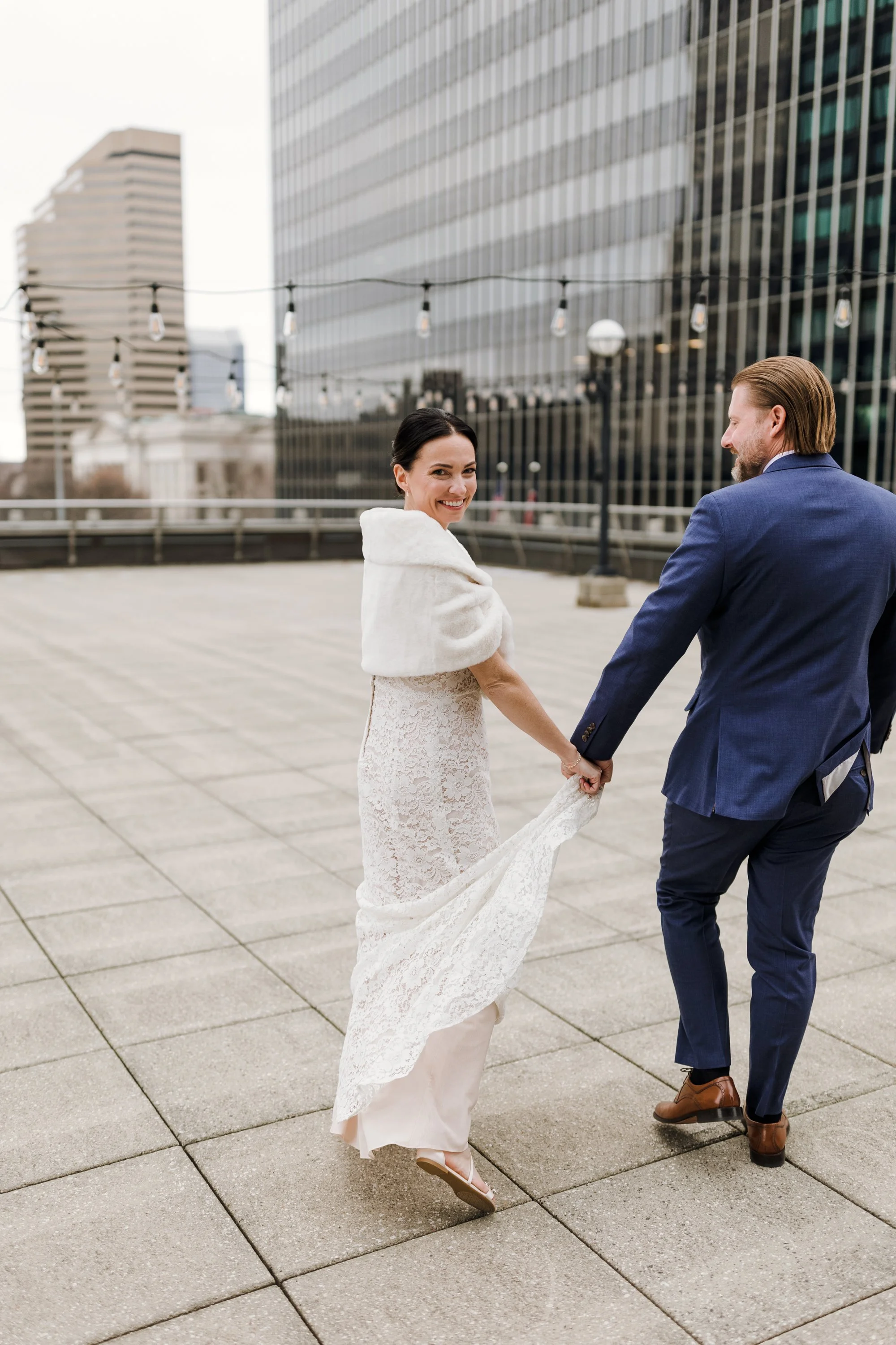 A bride in a white lace wedding dress and a white shawl holding hands with a groom in a blue suit on a rooftop in an urban setting.