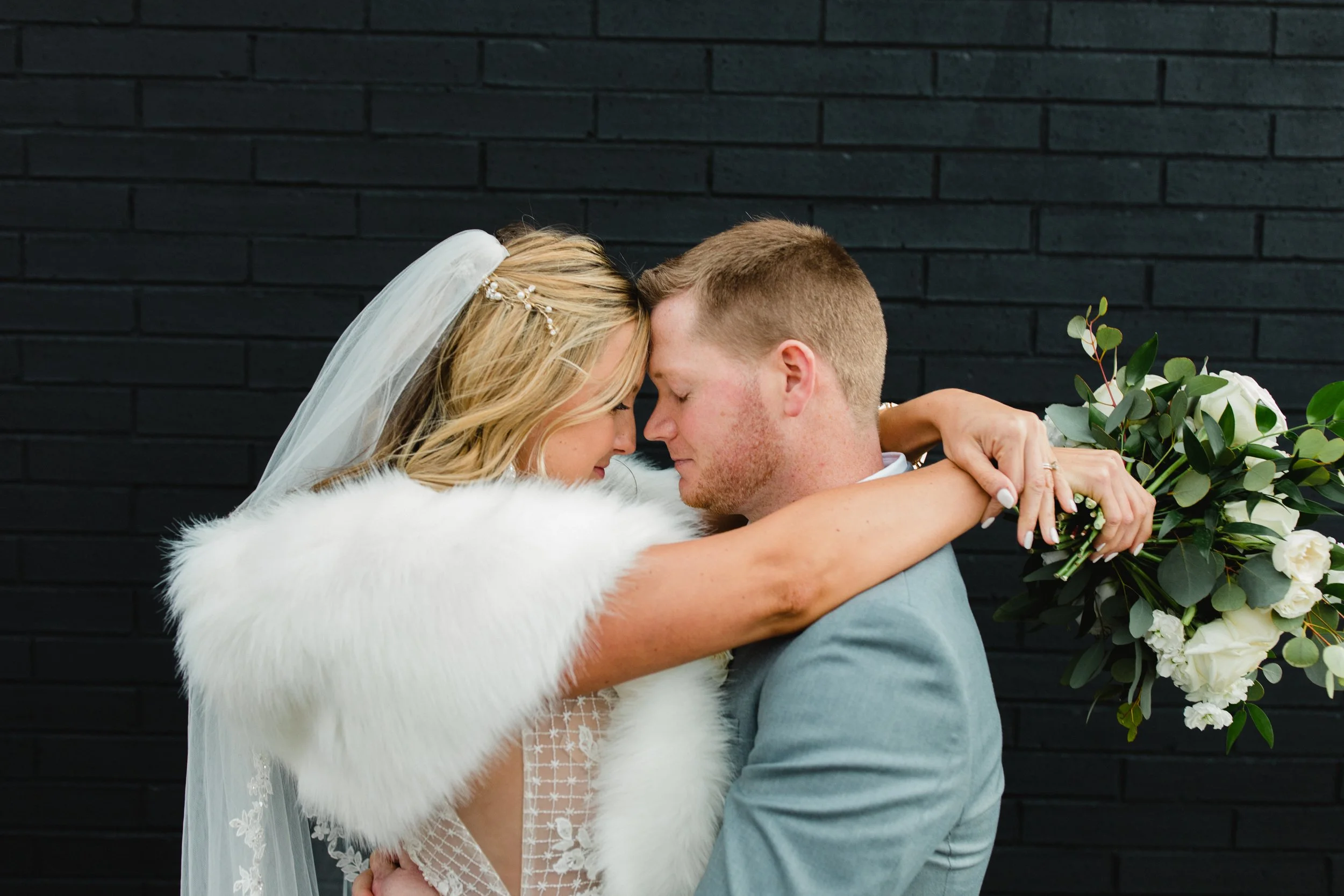 A bride and groom embrace with foreheads touching against a black brick wall. The bride wears a white veil, a white faux fur shawl, and holds a bouquet of white flowers and greenery. The groom wears a light gray suit.
