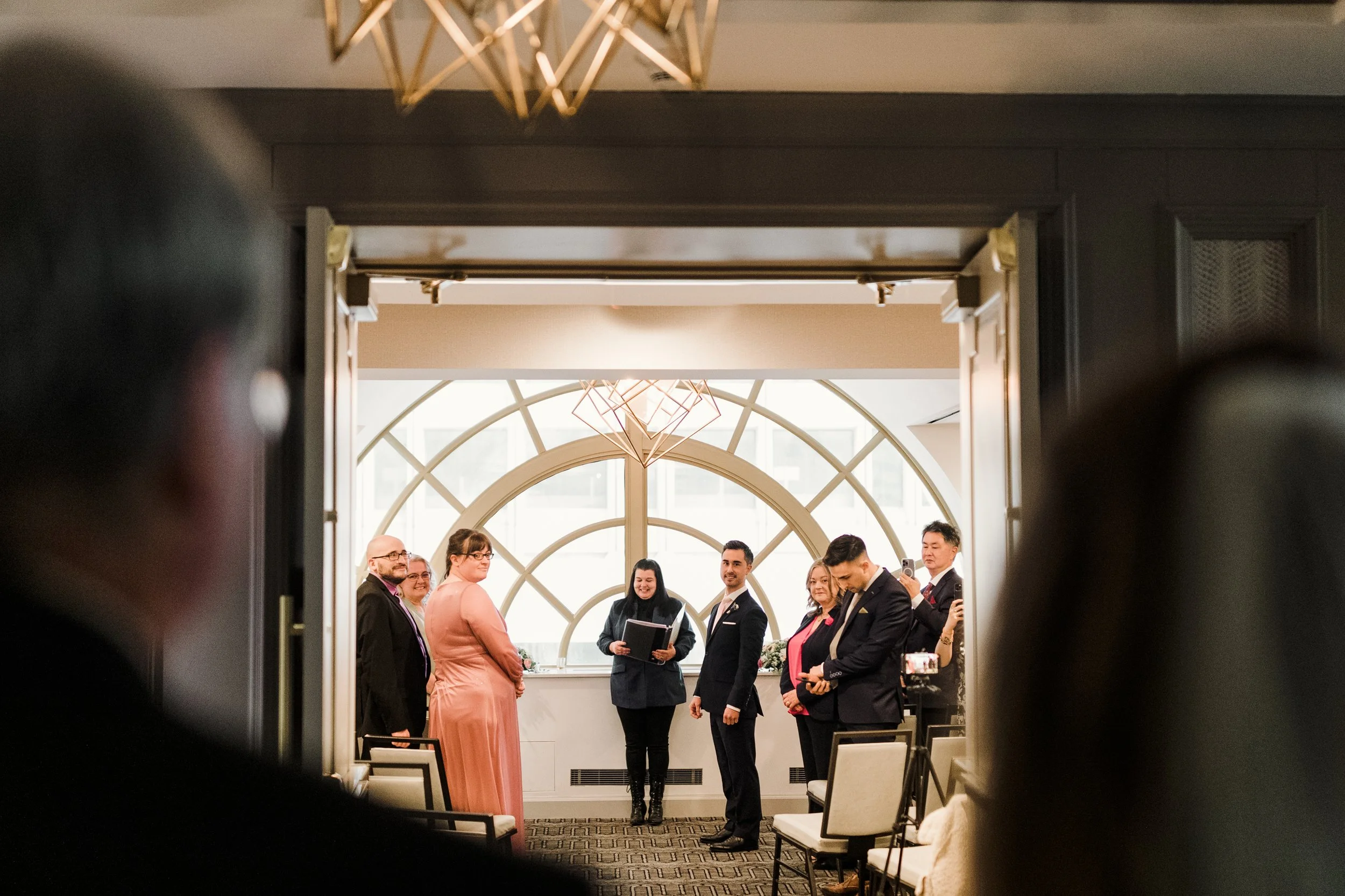 A group of people at a wedding ceremony, seen through a doorway, with some holding smartphones and others standing in front of a large window.