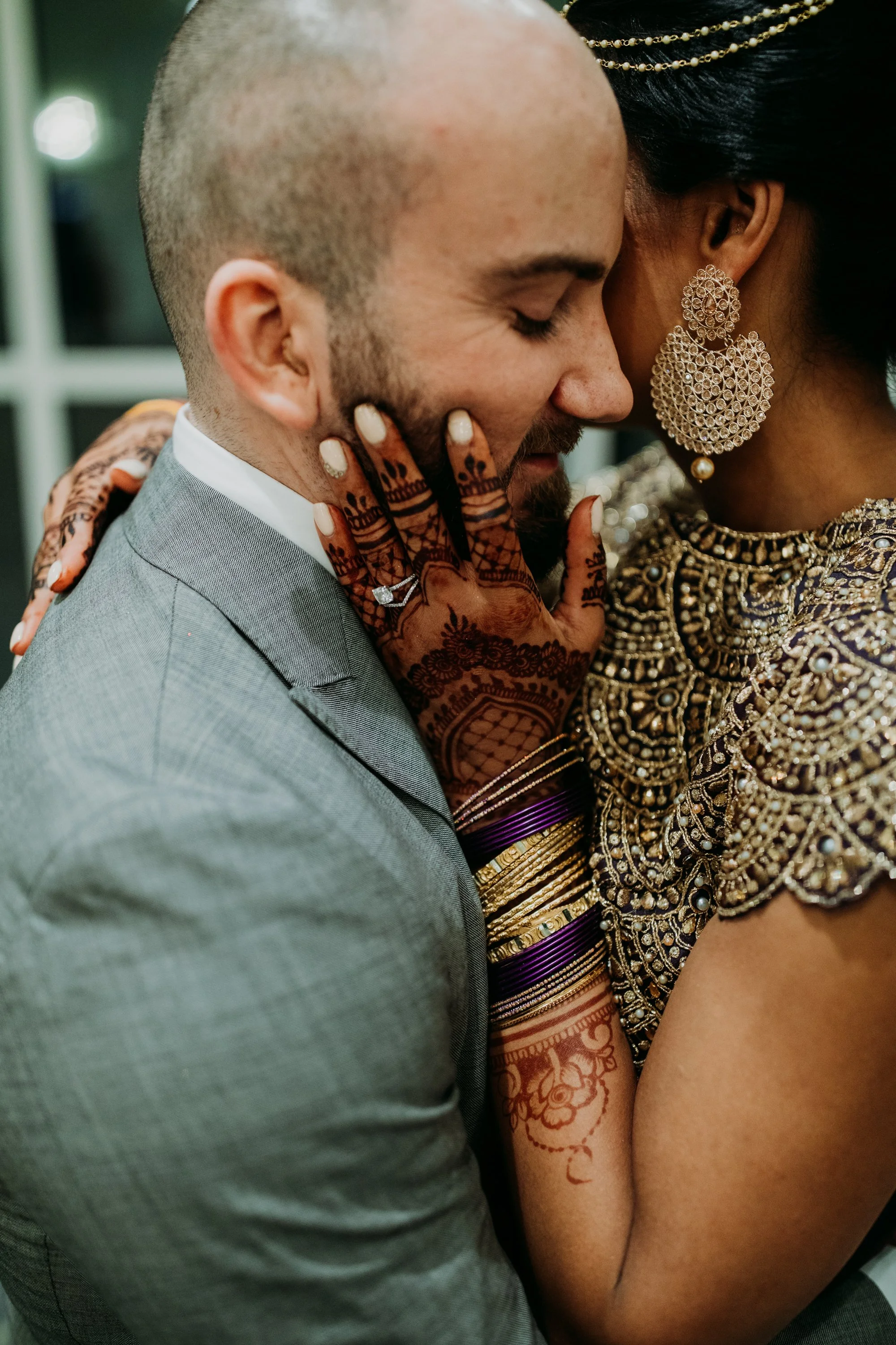 A couple hugging closely, with their foreheads and noses touching. The man is smiling, wearing a gray suit, while the woman is dressed in traditional Indian attire with detailed jewelry, and has henna designs on her arm.