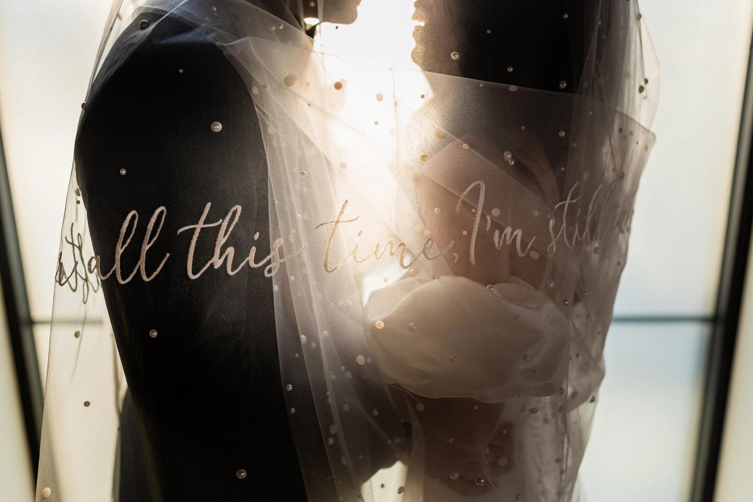 A bride and groom standing close together, united under a white veil with the text 'All this time, I'm still' written on it, backlit by sunlight streaming through a window.