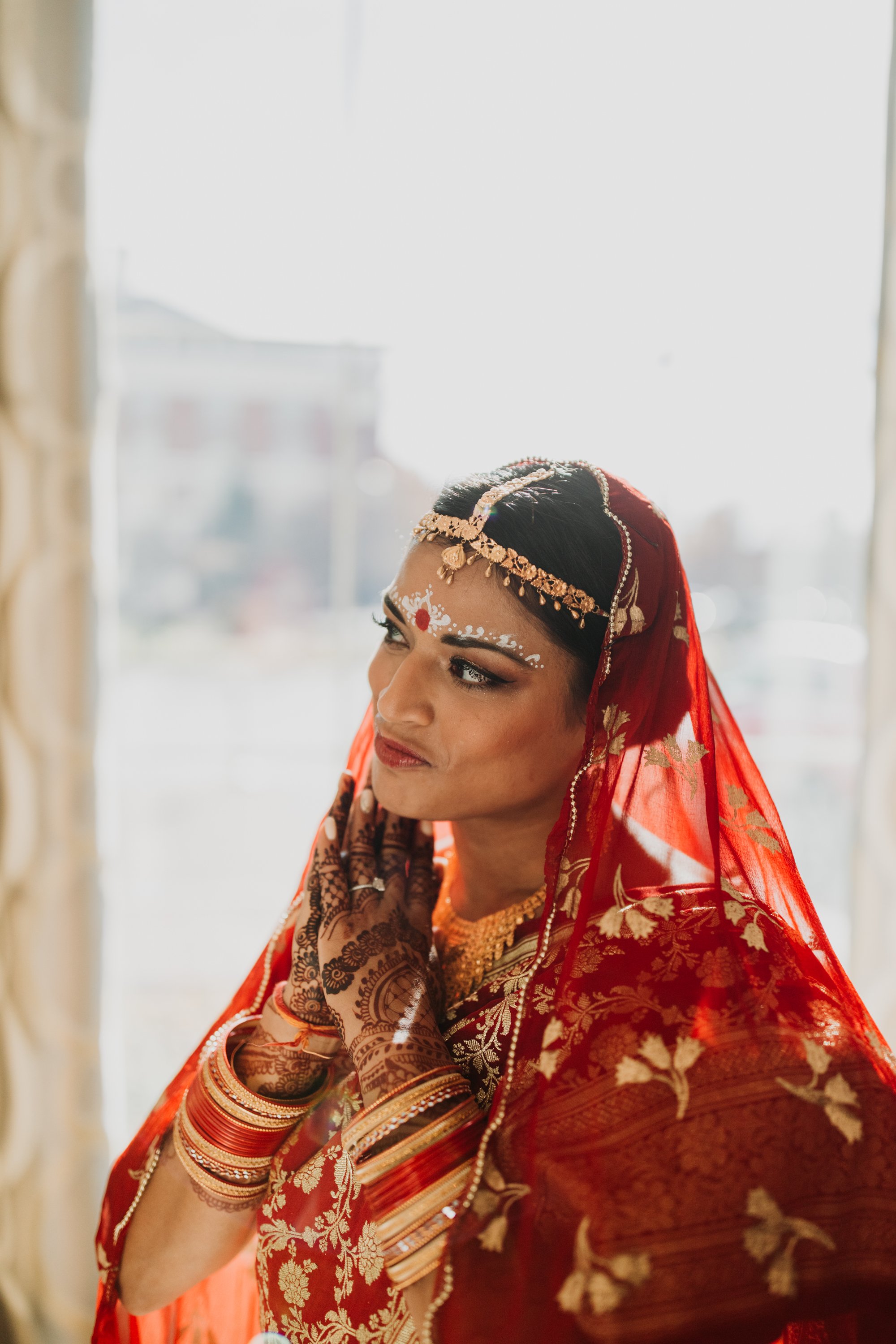 A woman dressed in traditional Indian wedding attire, wearing a red saree with gold embroidery, gold jewelry, and henna on her hands, standing near a window with sunlight.