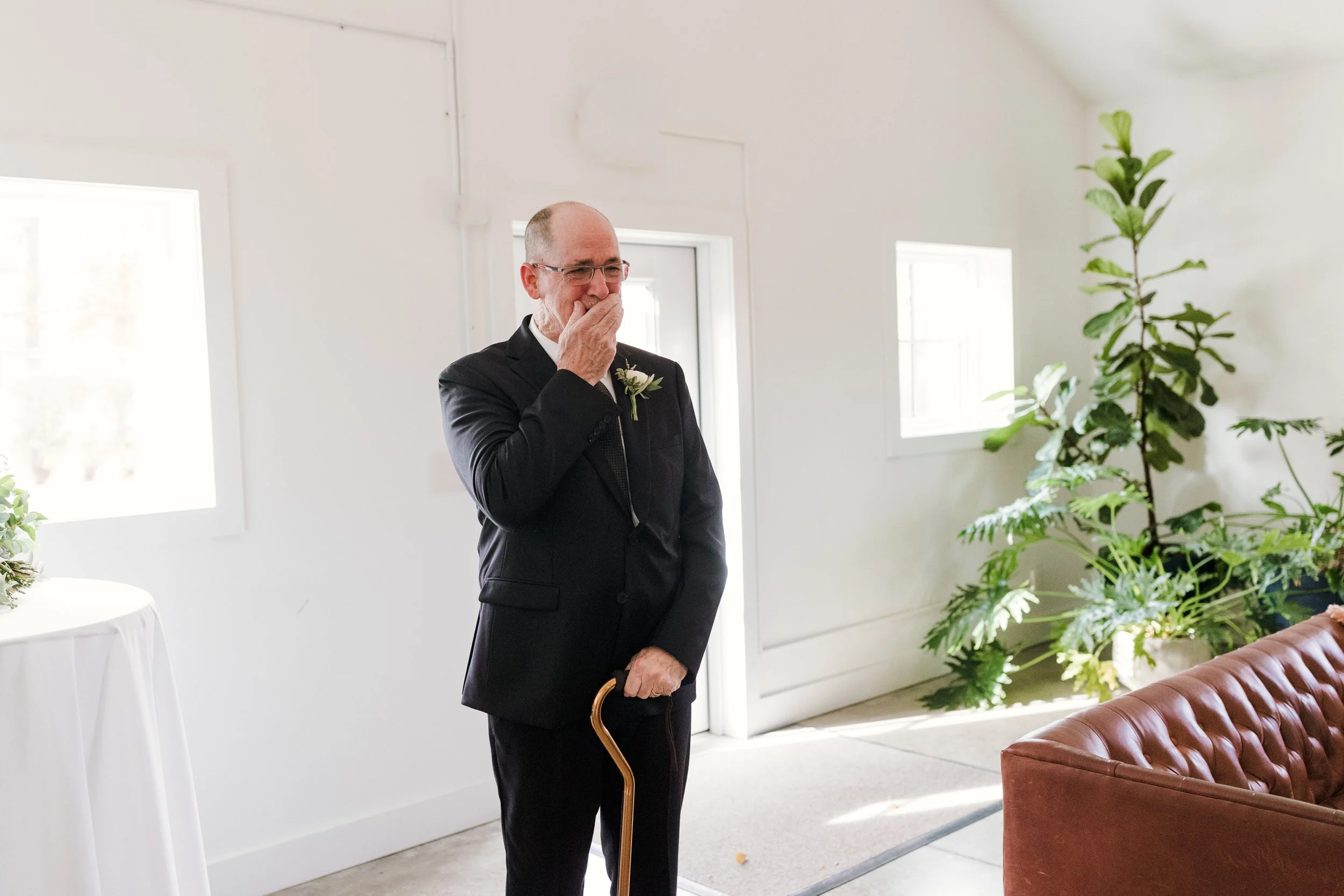 An elderly man in a black suit with a cane covers his mouth with his hand, appearing emotional, in a brightly lit room with white walls, windows, and potted plants.