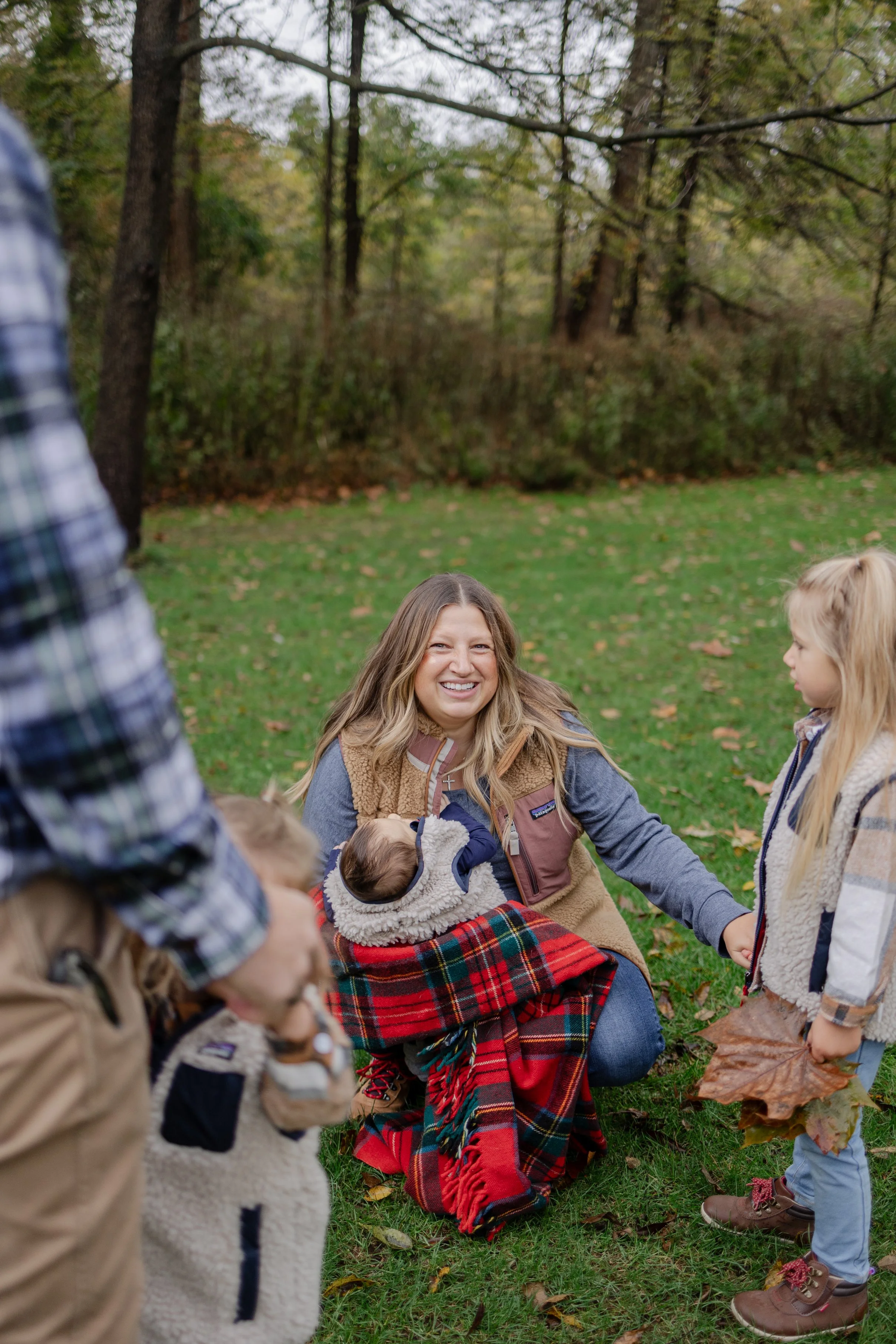 A woman smiling and holding a baby, with children around her in a park with trees and fallen leaves.