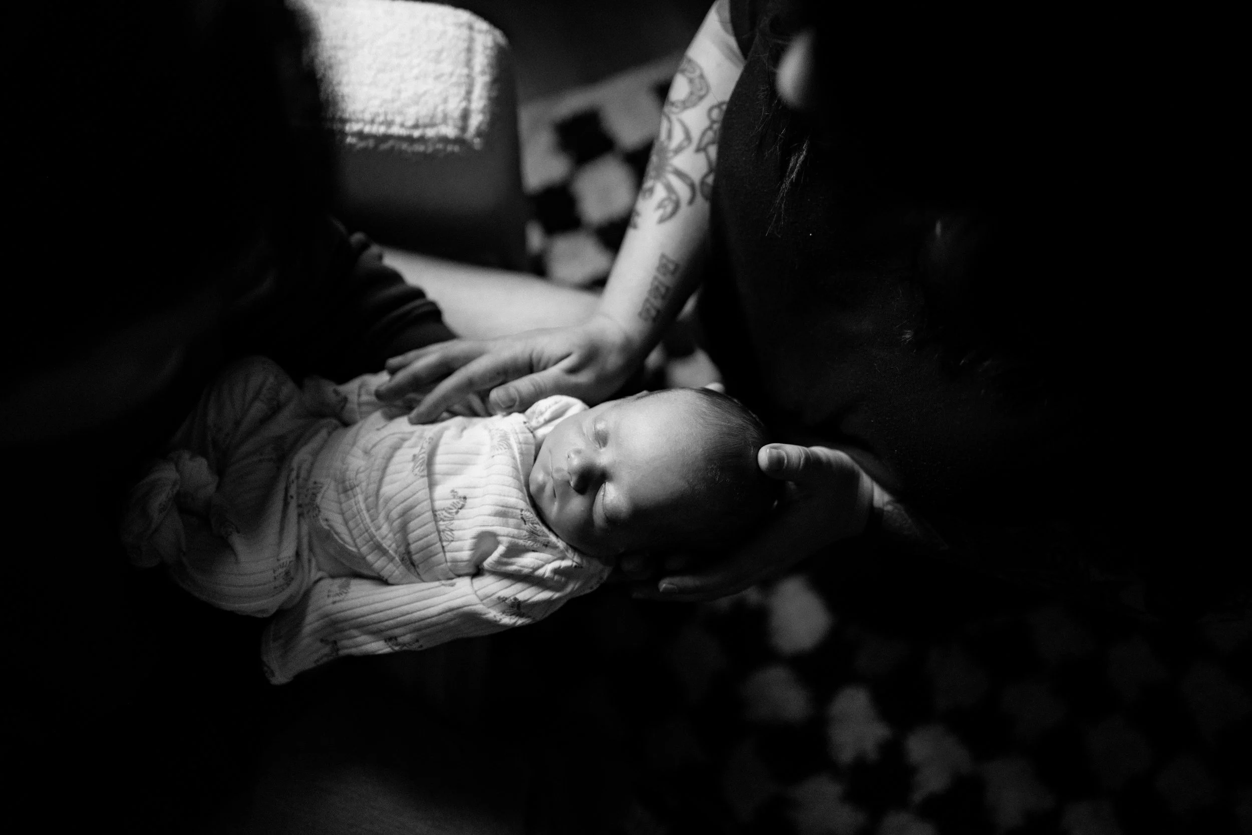 Black and white photo of a sleeping baby being gently touched on the head and chest by two adults, one on each side, in a caring and intimate moment.