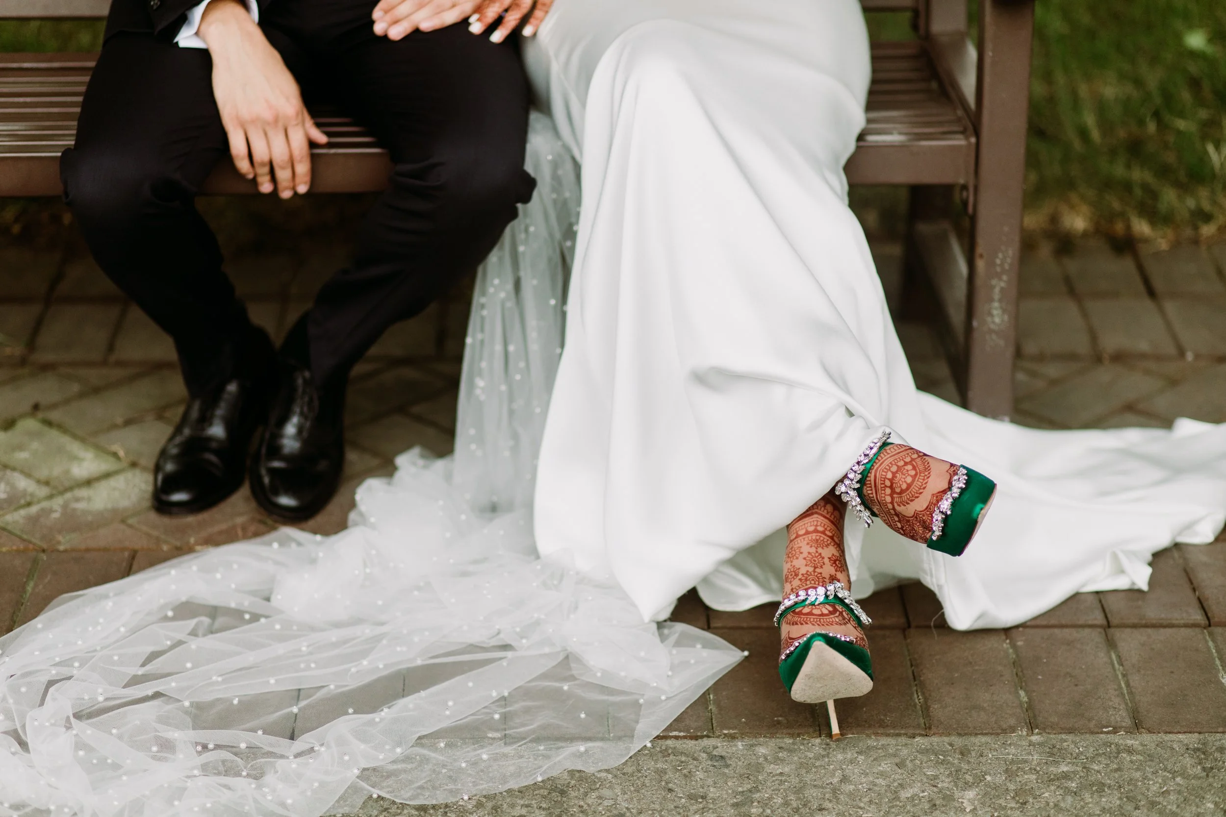 Close-up of a bride and groom sitting on a park bench, showing part of the bride's white dress and ornate green and gold high heels, with the groom dressed in black suit and shoes.