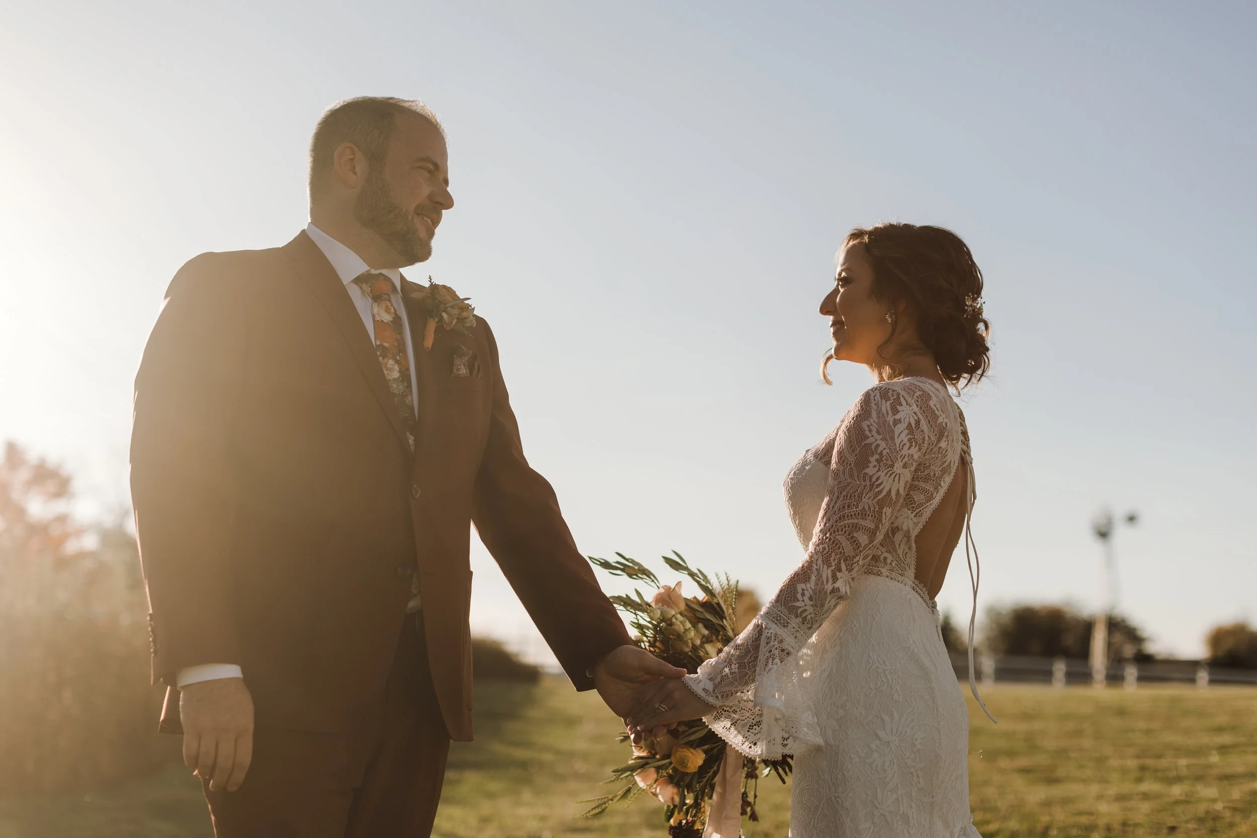 A bride and groom holding hands and smiling at each other outdoors during sunset, with the groom in a brown suit and the bride in a lace wedding dress, holding a bouquet of flowers.
