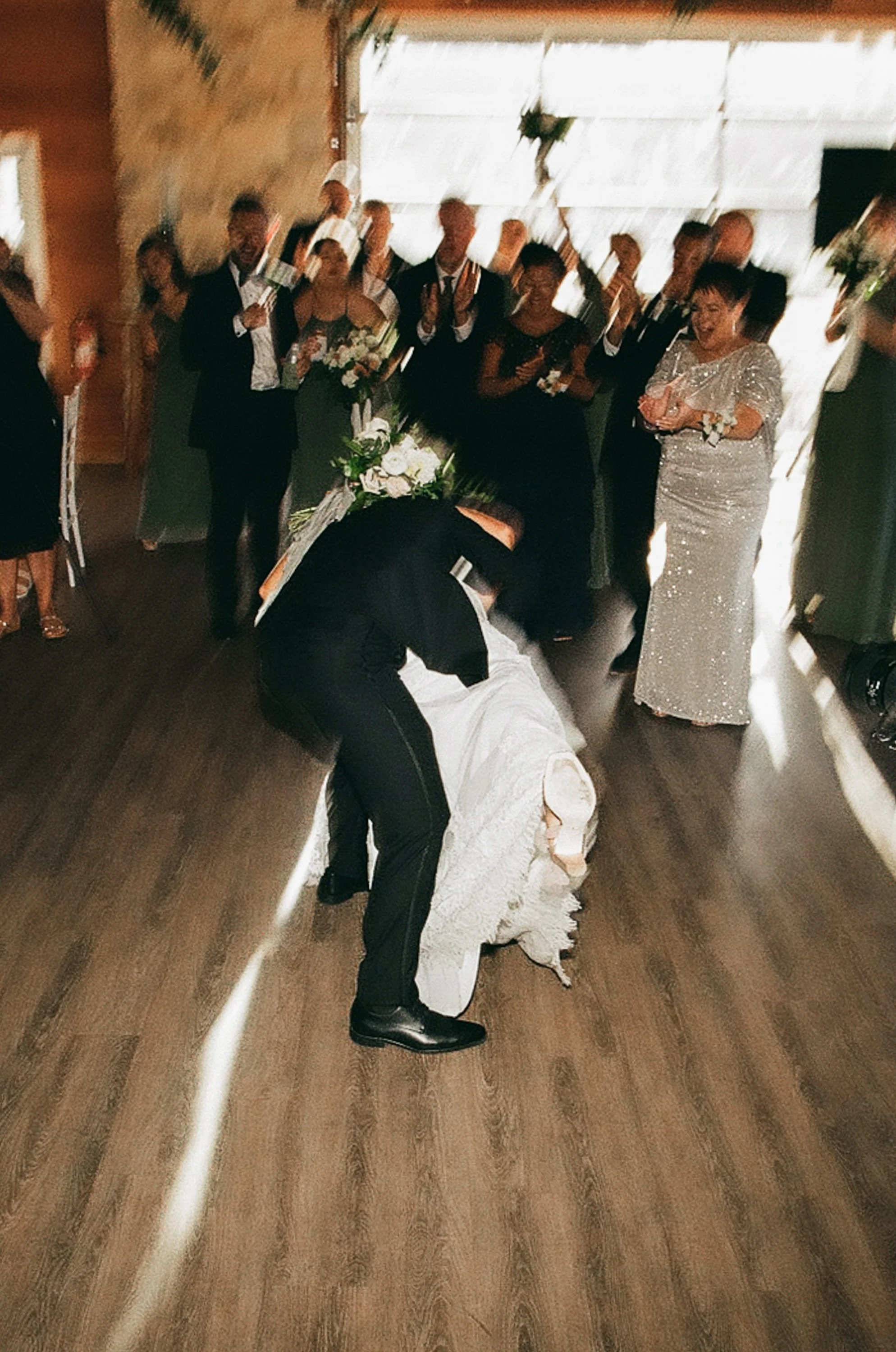 A wedding reception scene with a groom kissing the bride on the floor, surrounded by guests clapping and smiling.