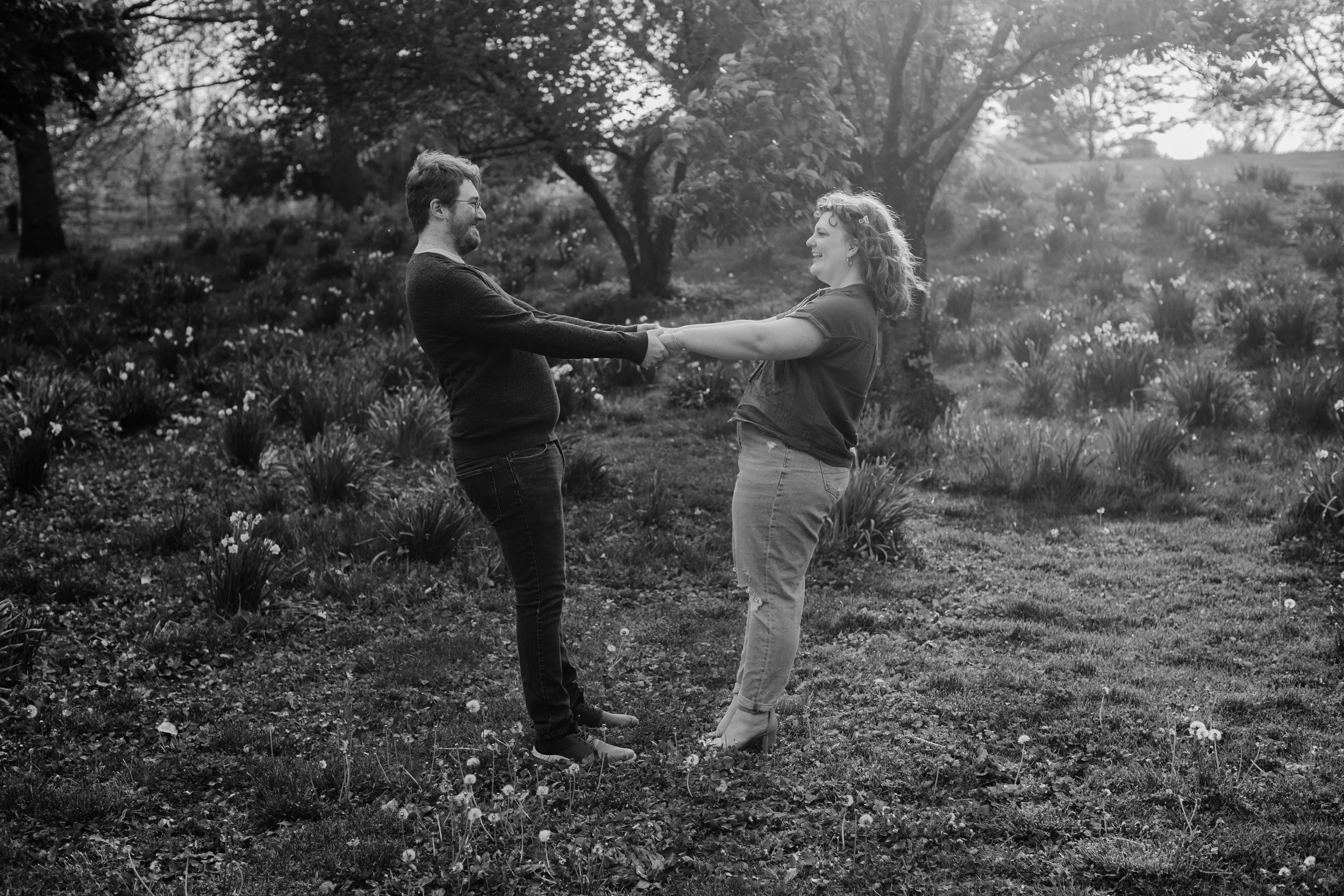 A black and white photo of a couple holding hands and smiling at each other in a garden or park with trees and flowers.