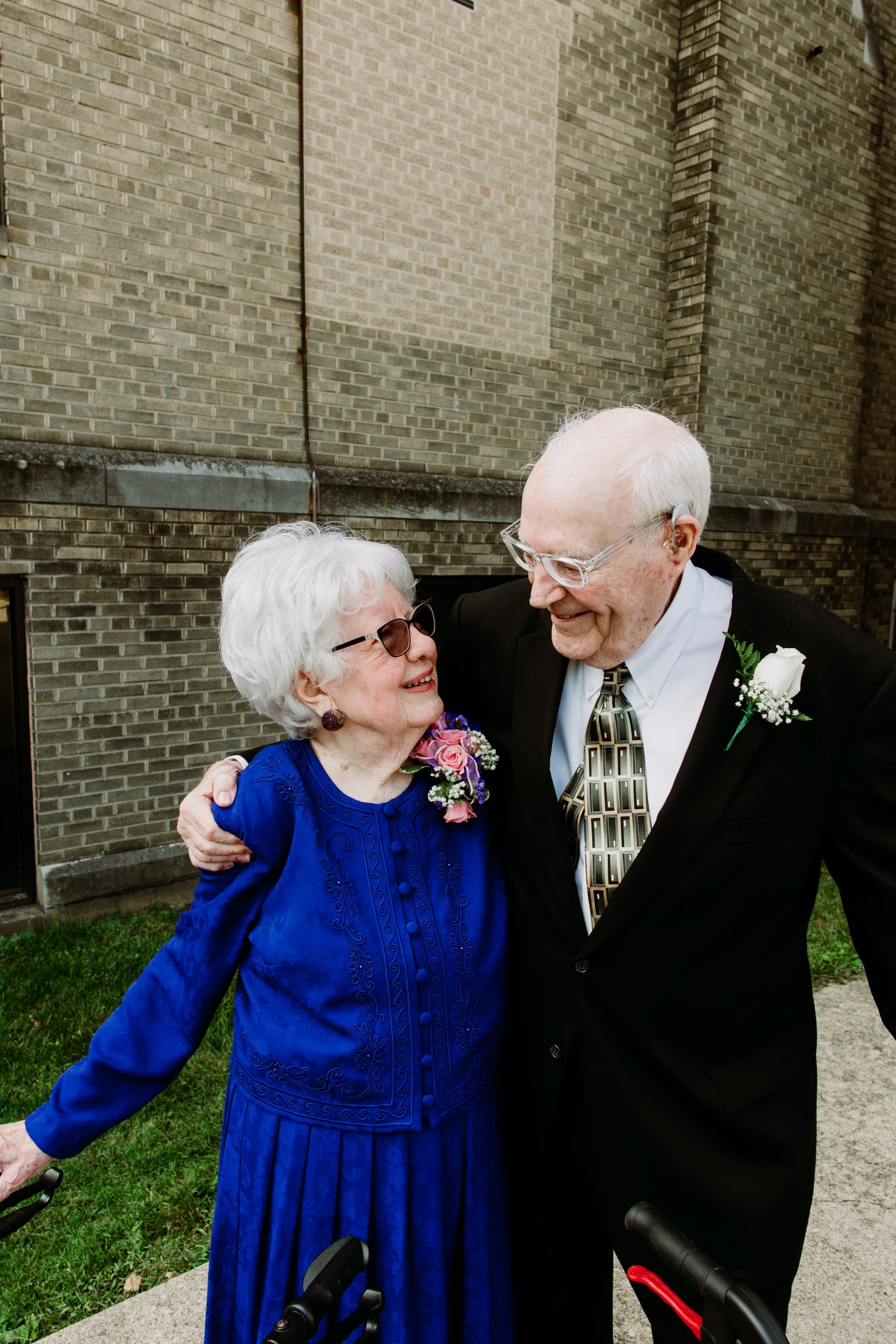 An elderly woman and man smiling and embracing outside near a brick building.