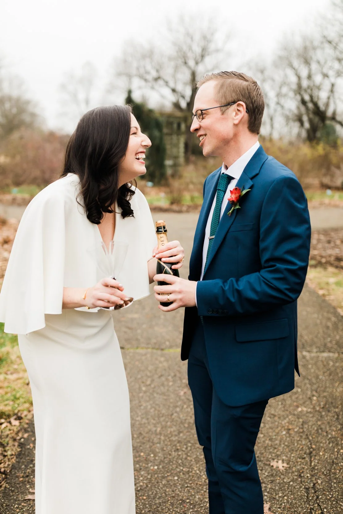 A couple dressed in wedding attire, celebrating outdoors with champagne in hand, smiling and looking at each other against a backdrop of leafless trees and overcast sky.