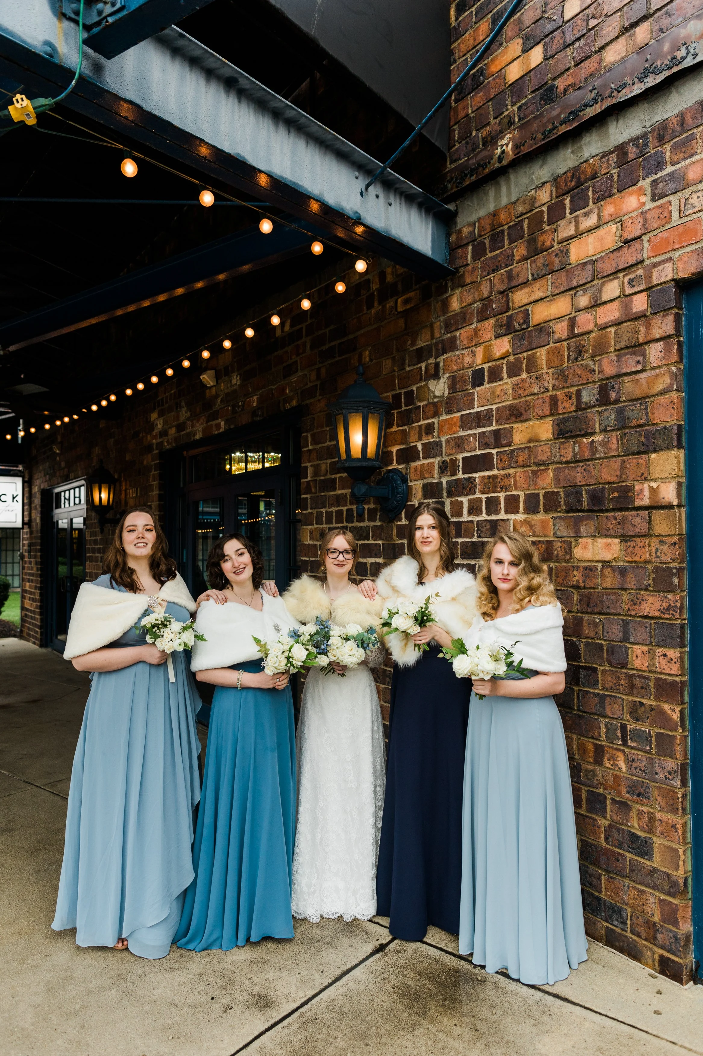 Group of five women in bridesmaid dresses and a bride in a white gown, standing outside near a brick building, holding bouquets of white flowers, with string lights overhead.