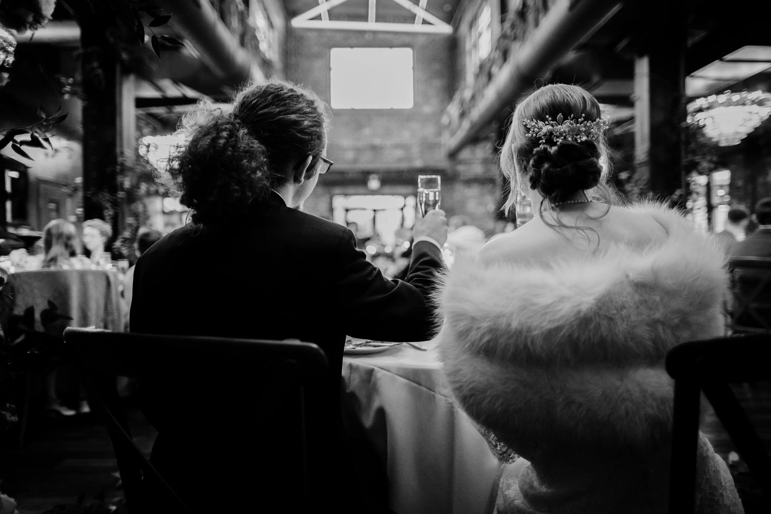 A black and white photo of a couple sitting at a table during a celebration, with the person on the left raising a glass, and the person on the right wearing a floral headpiece and a fur stole, inside a spacious, industrial-style venue with high ceil