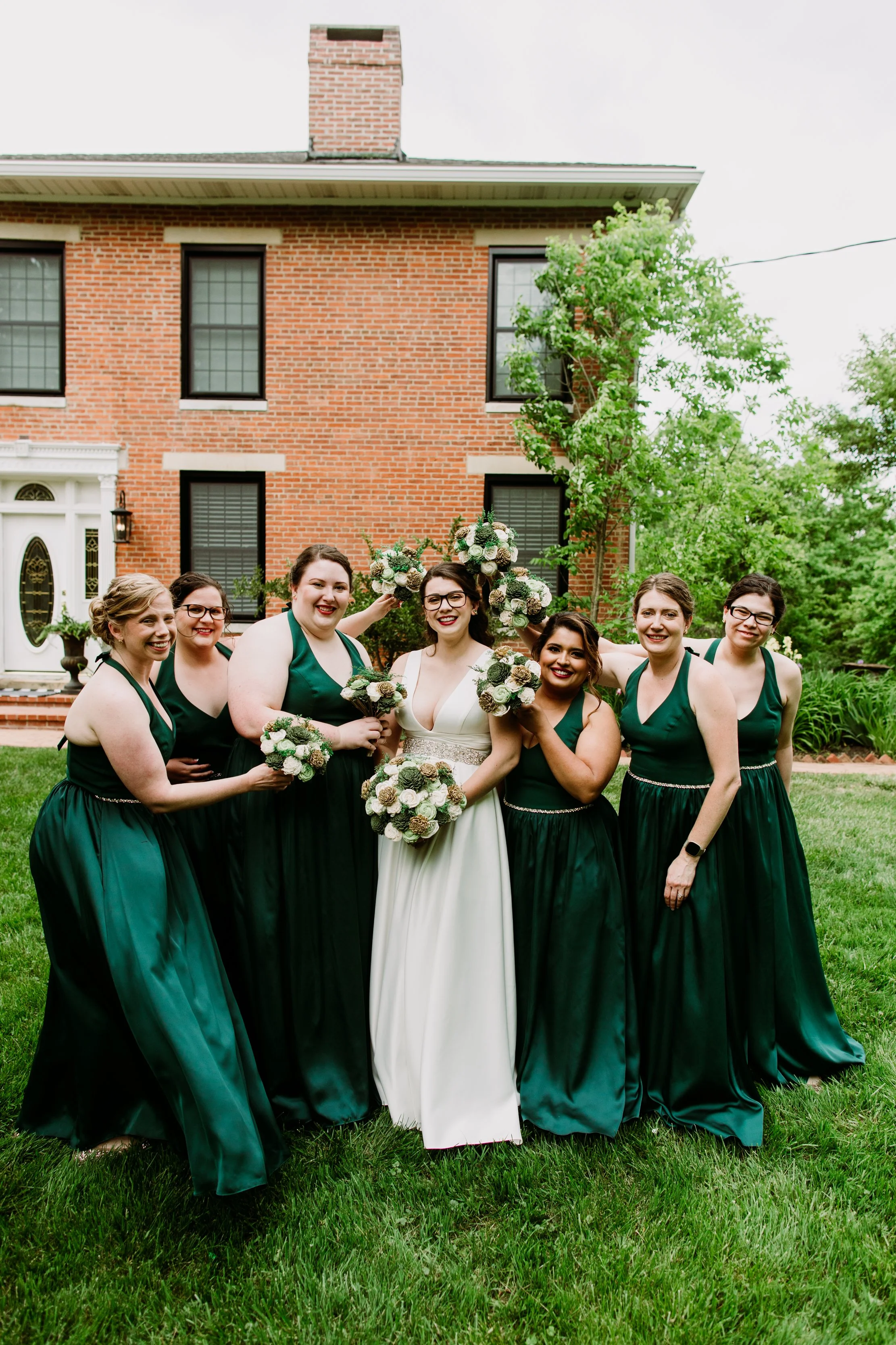 Group of women dressed in green bridesmaid dresses and a bride in a white wedding gown, holding bouquets, standing outside in front of a brick house with greenery and trees.