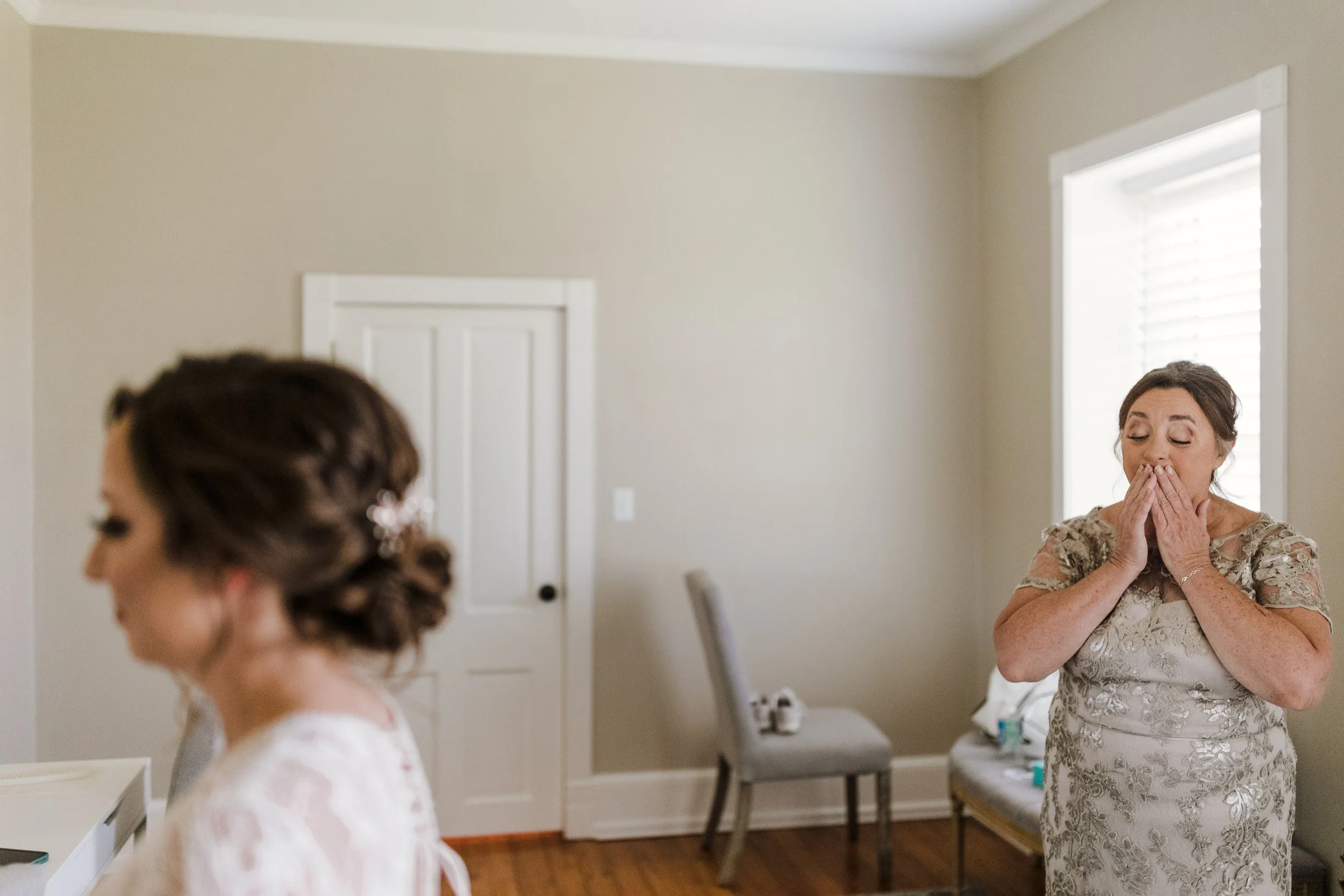 A woman in a wedding dress with her hair in an updo, standing in a room with light-colored walls, and another woman in a lace dress with her hands covering her mouth, appearing emotional, standing near a window with closed blinds.