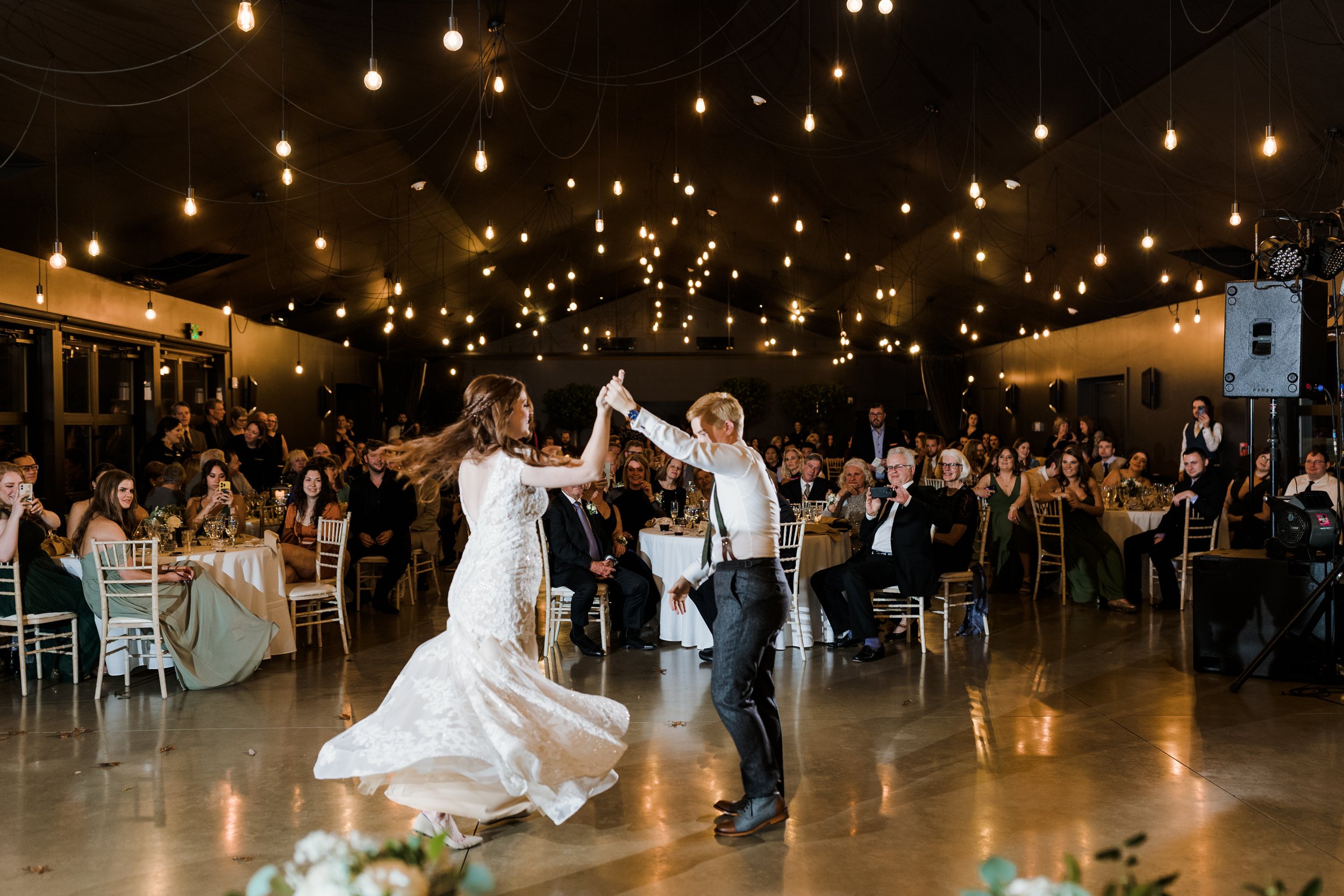 A bride and groom dancing at their wedding reception, surrounded by seated guests in a decorated venue with hanging lights.