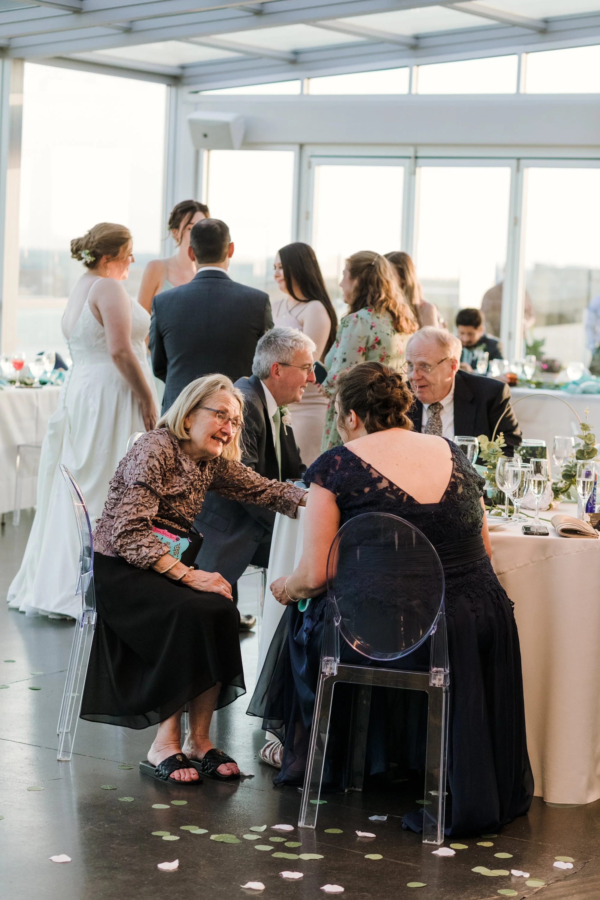 People gathered at a wedding reception, with a bride and groom standing and friends and family sitting and talking at a table near large windows.