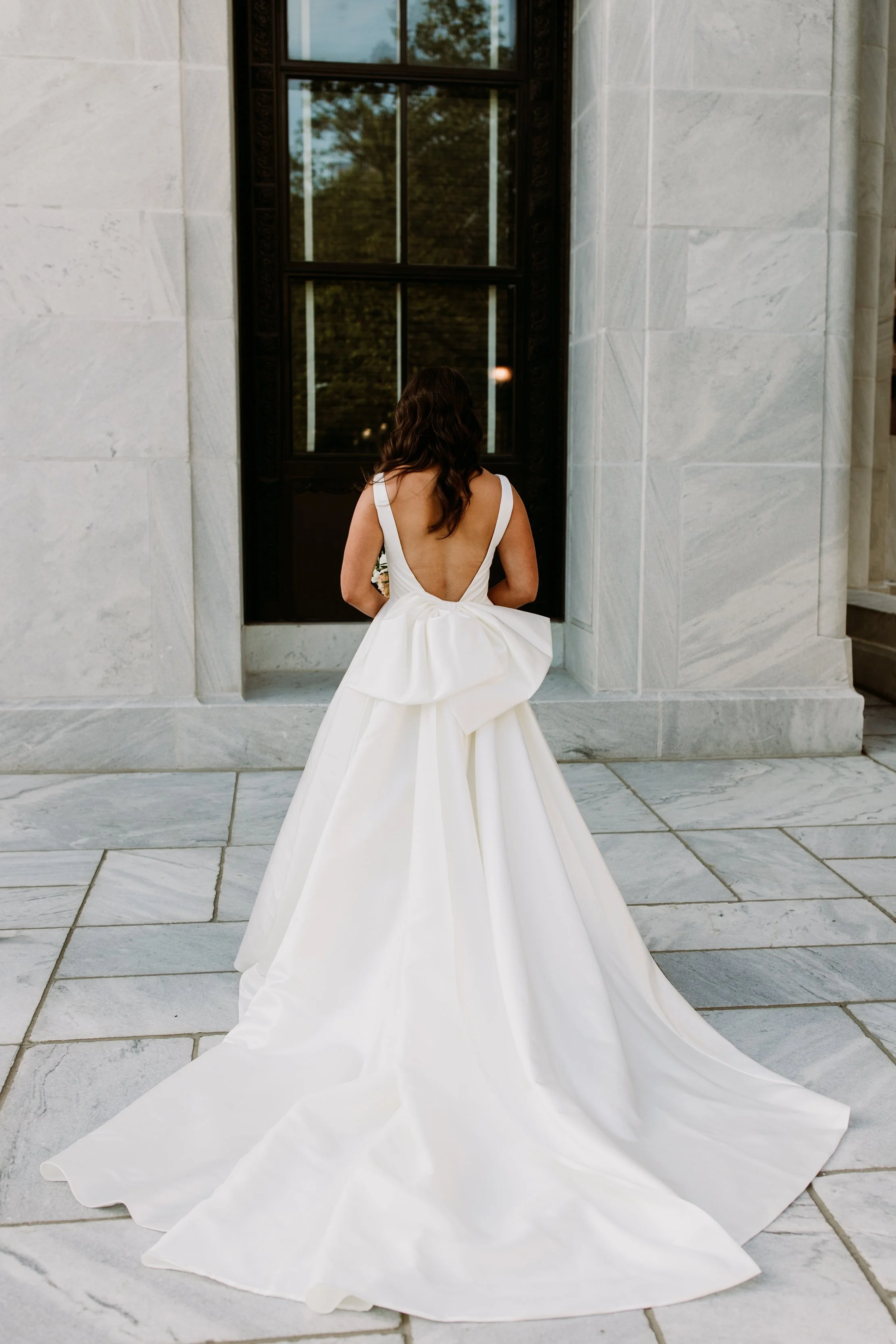Woman in a white wedding dress with an open back, standing in front of a tall, black-framed window on a marble building exterior.