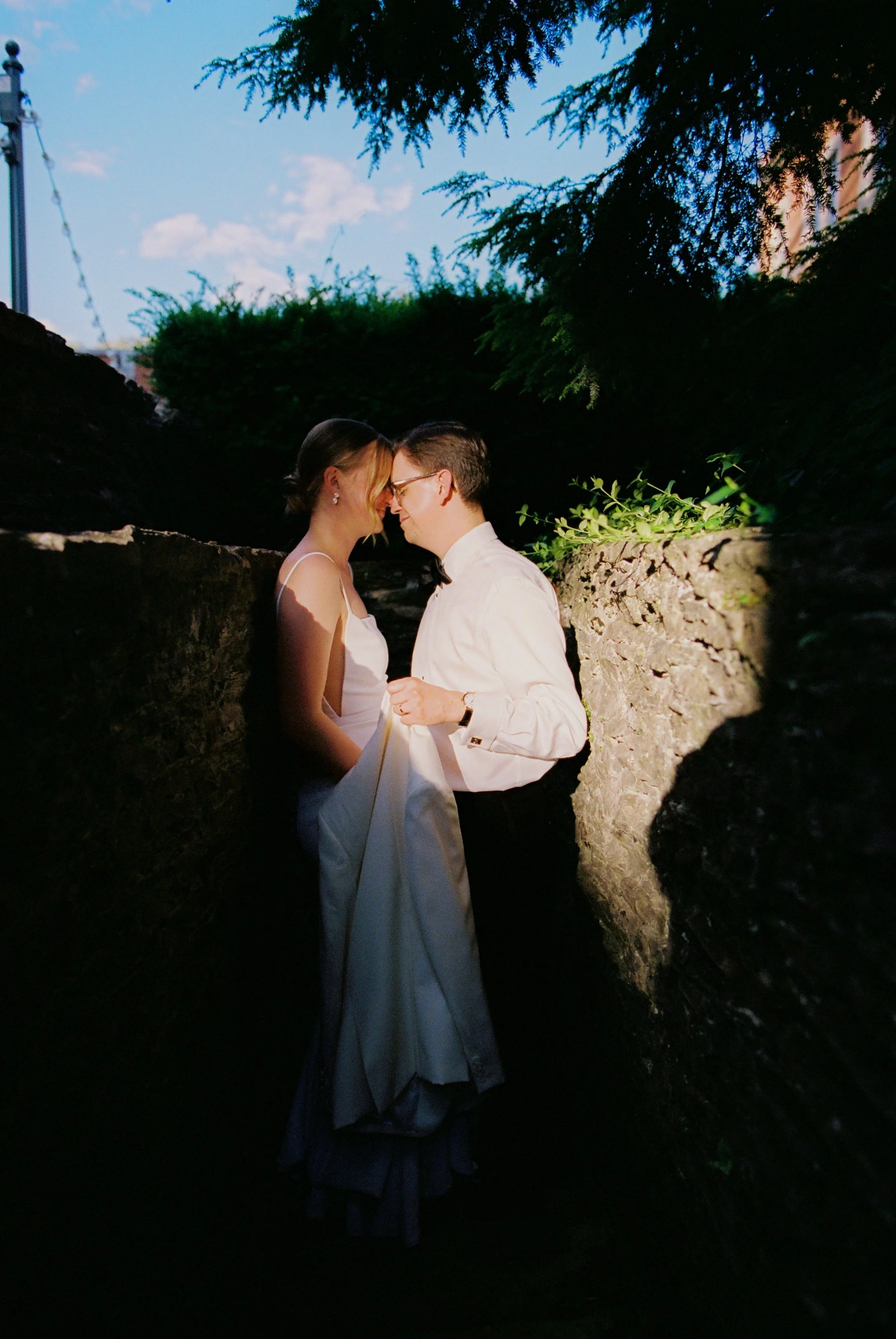 A couple stands close together in an intimate embrace, with foreheads touching and eyes closed, between two stone walls outdoors during daytime, with trees and blue sky in the background.