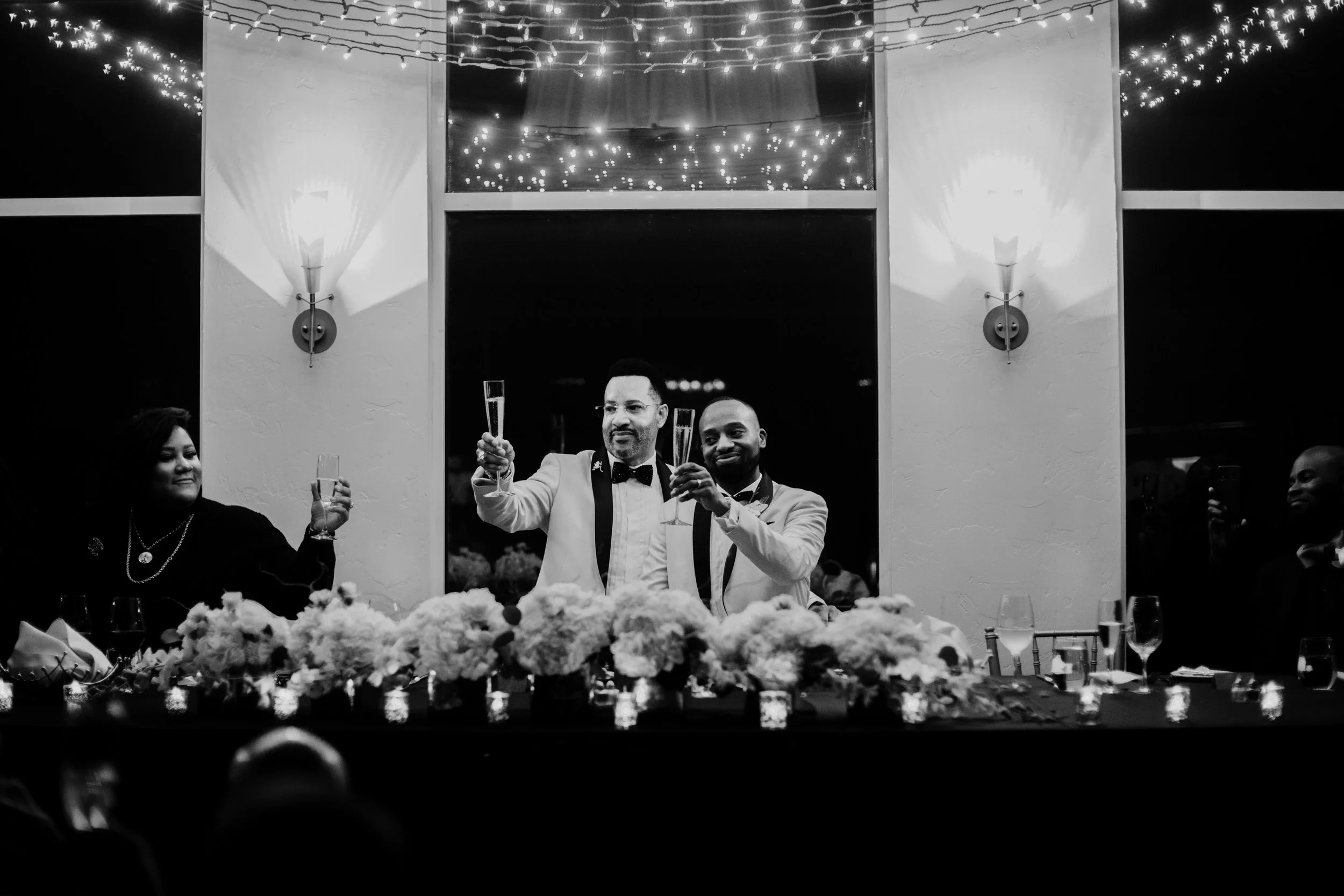 Black and white photo of a celebration with two men in tuxedos raising champagne glasses, surrounded by women and guests, with floral decorations and string lights in the background.
