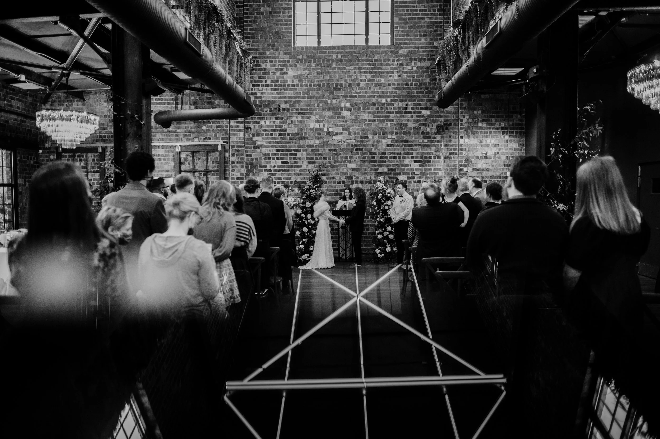 A black and white photo of a wedding ceremony inside a venue with exposed brick walls and industrial decor. The couple is standing at the altar facing each other, surrounded by floral arrangements. Guests are standing and watching the ceremony.