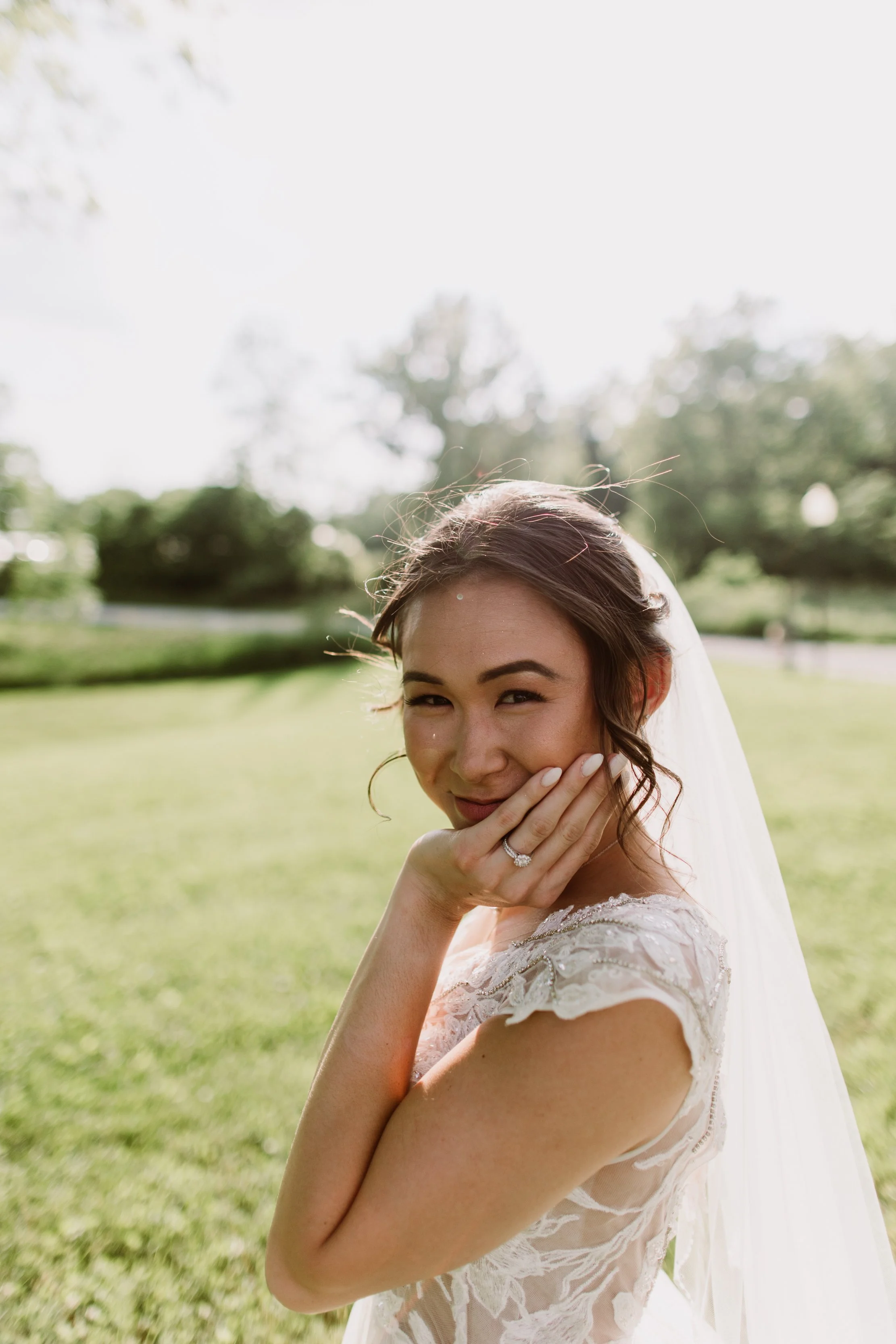 A bride standing outdoors on a sunny day, smiling and touching her face with one hand, wearing a wedding dress with lace detail and a veil.