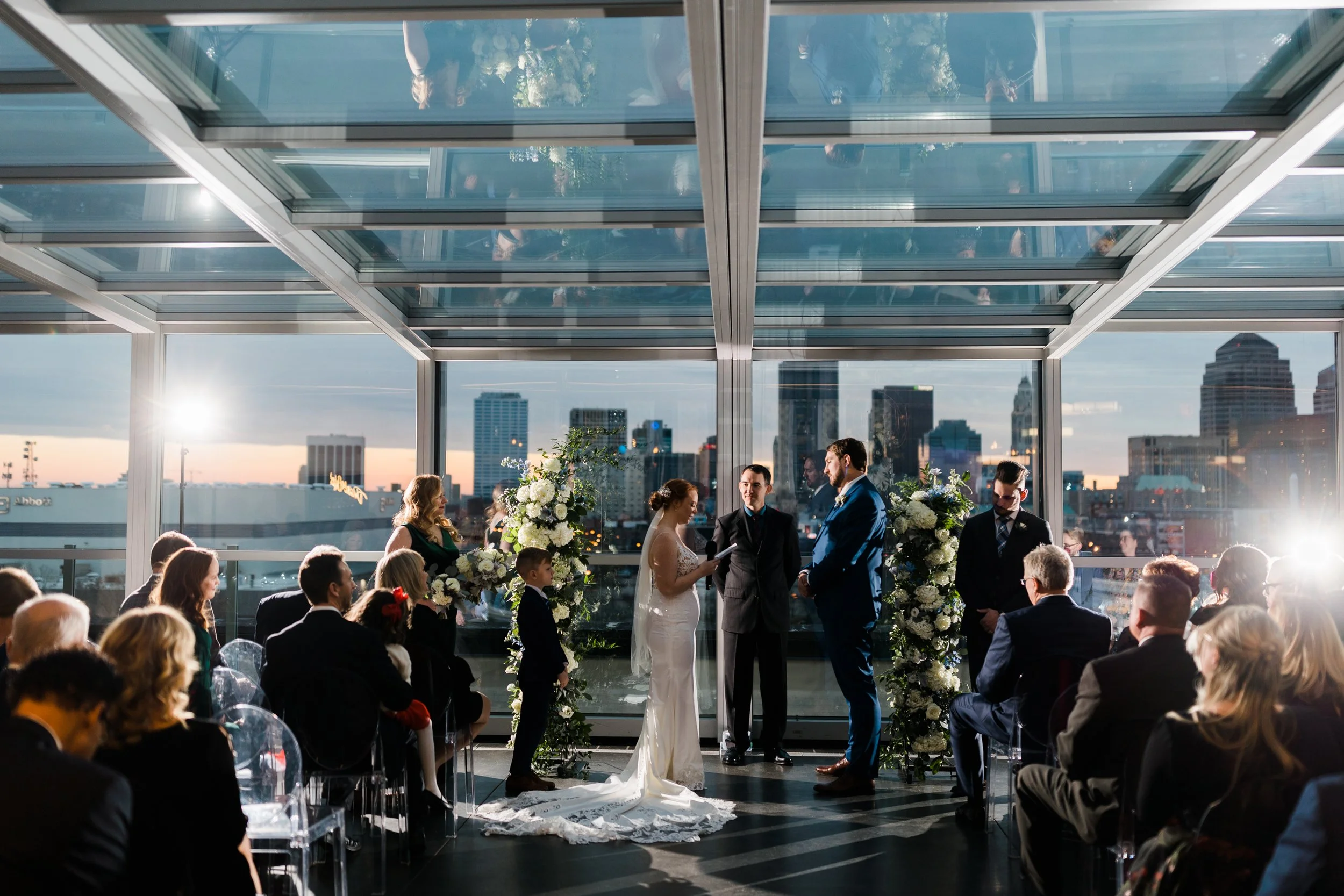 A wedding ceremony occurs in a modern glass venue with city skyline views at sunset, with the bride and groom exchanging vows, surrounded by floral arrangements and guests seated on transparent chairs.