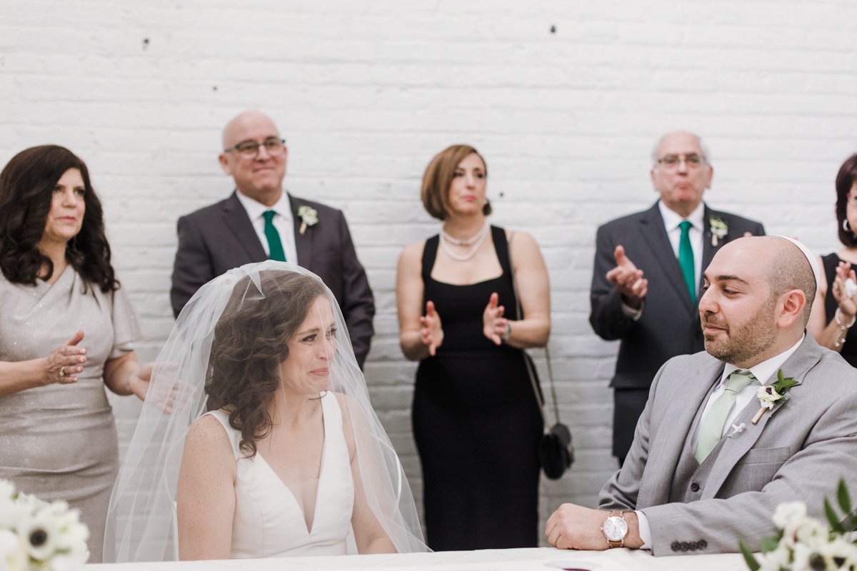 A bride and groom sit at a table during their wedding ceremony, with family and friends clapping and watching in the background.