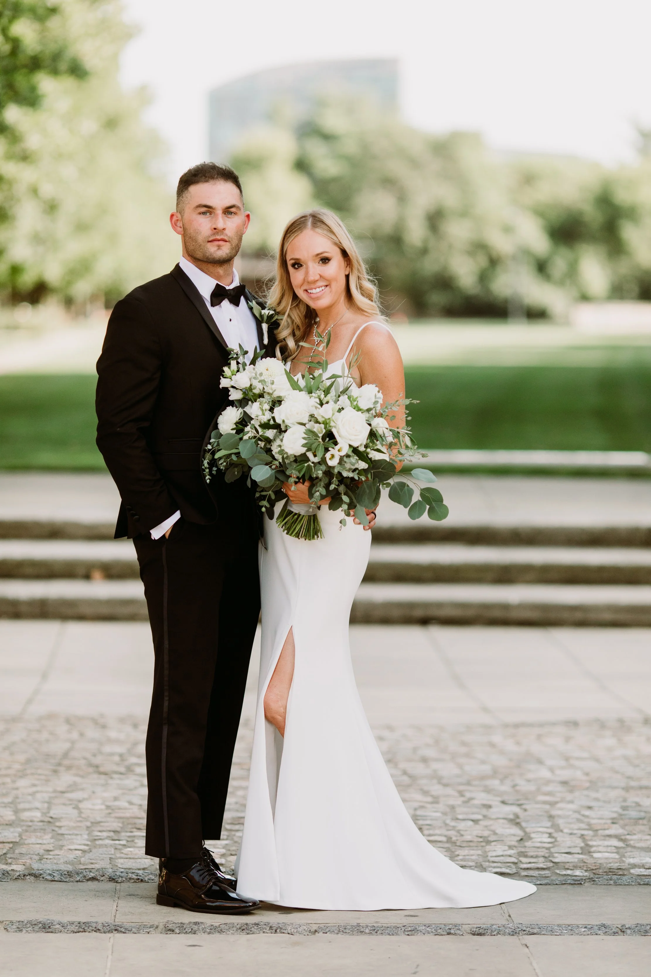 A bride and groom standing outdoors on a wedding day, with the bride holding a large bouquet of white flowers and greenery, both dressed elegantly with a park and trees in the background.