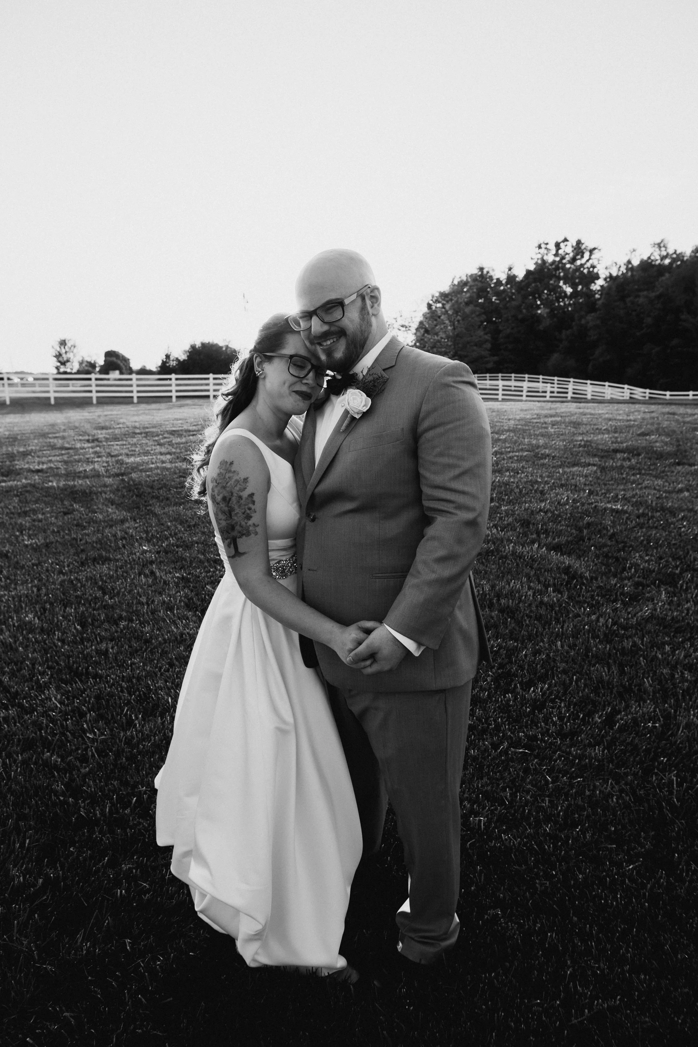 Black and white photo of a bride and groom hugging outdoors on a grassy field with a white fence and trees in the background, smiling and holding hands.