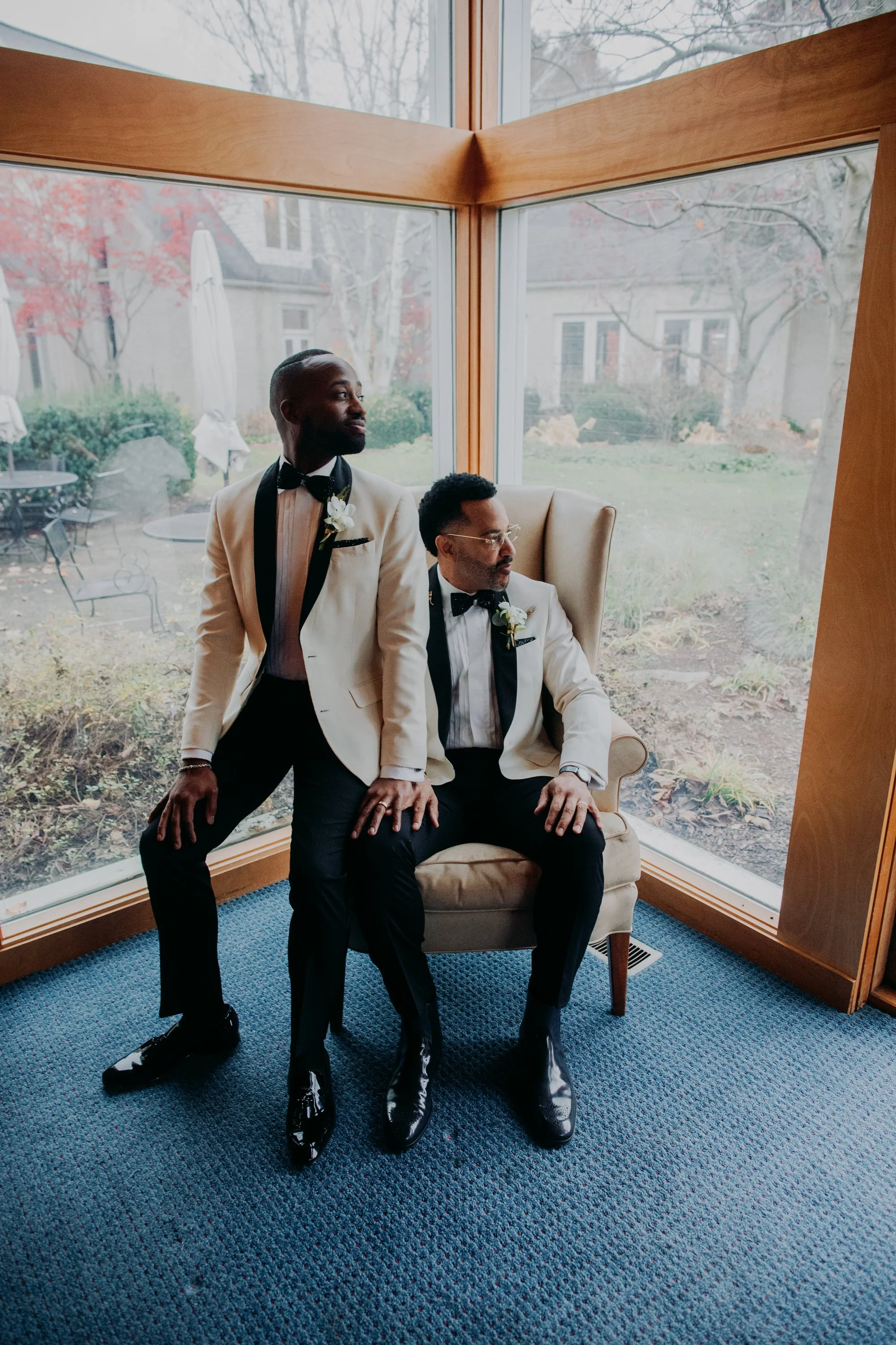 Two men dressed in tuxedos, one seated in a beige armchair and the other standing beside him, are inside a room with large windows showing the outdoors with trees and an outdoor patio.
